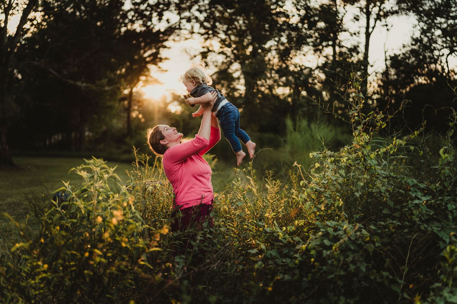 Parent lifts child joyfully in wildflower field at sunset with golden light filtering through trees.