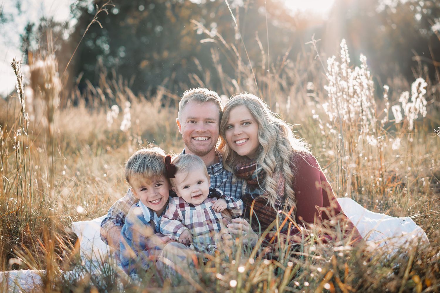 A family sits together on a blanket in a sun-drenched field of tall grass.