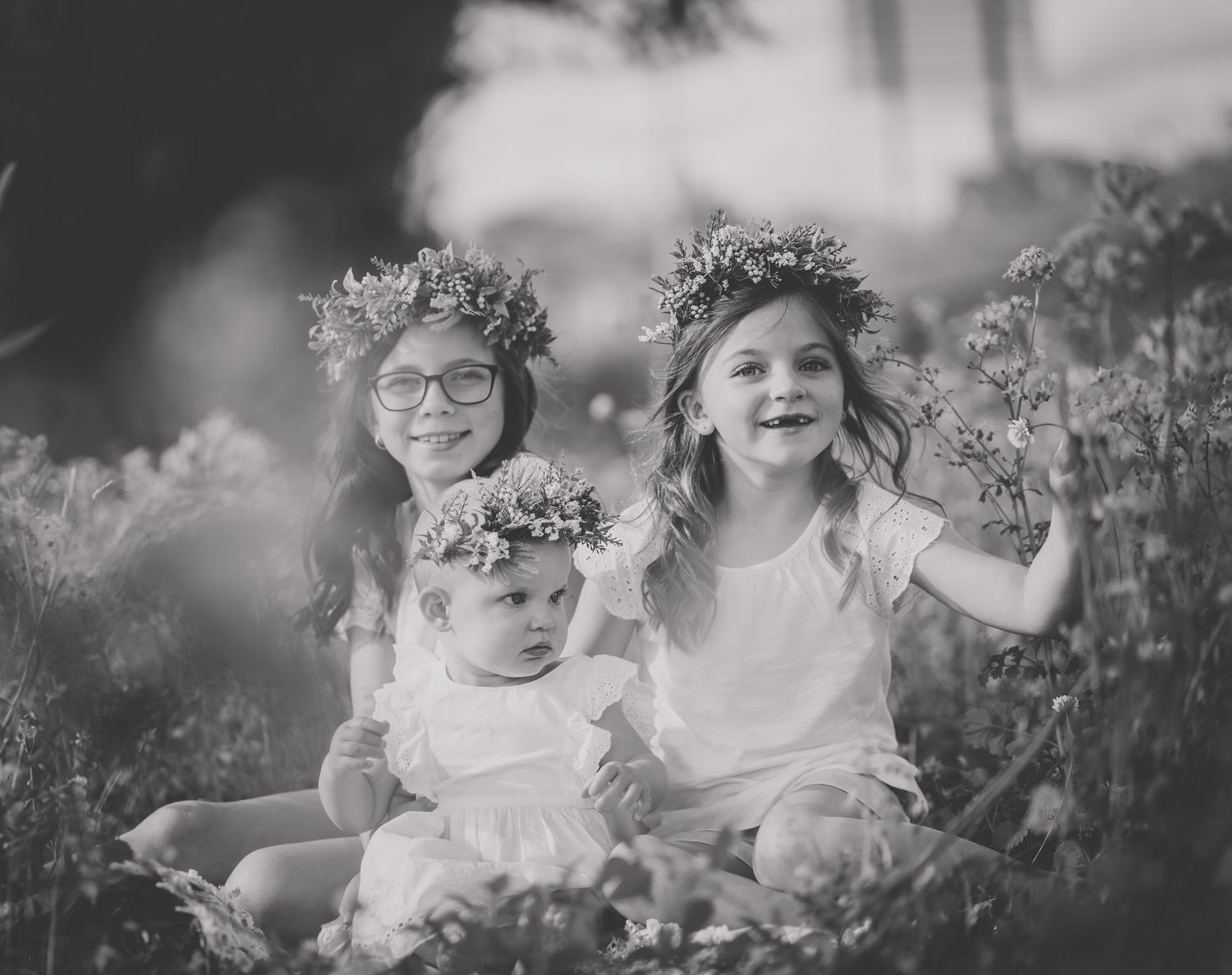 Black and white capture of three siblings wearing floral crowns sitting together outdoors.