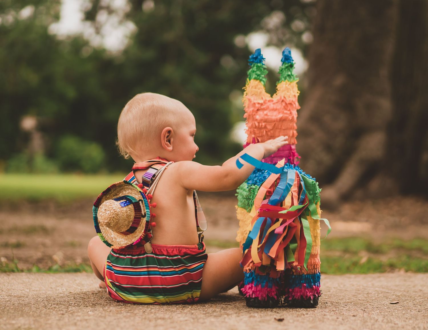 Baby in striped outfit sits beside colorful pinata on outdoor path.