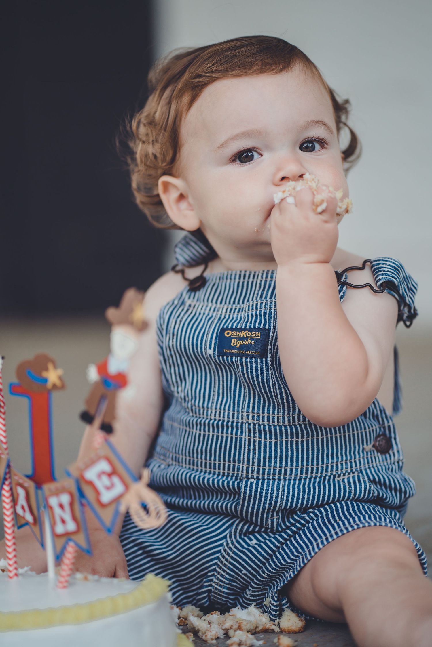 Baby in denim overalls enjoys birthday cake with messy frosting on face.