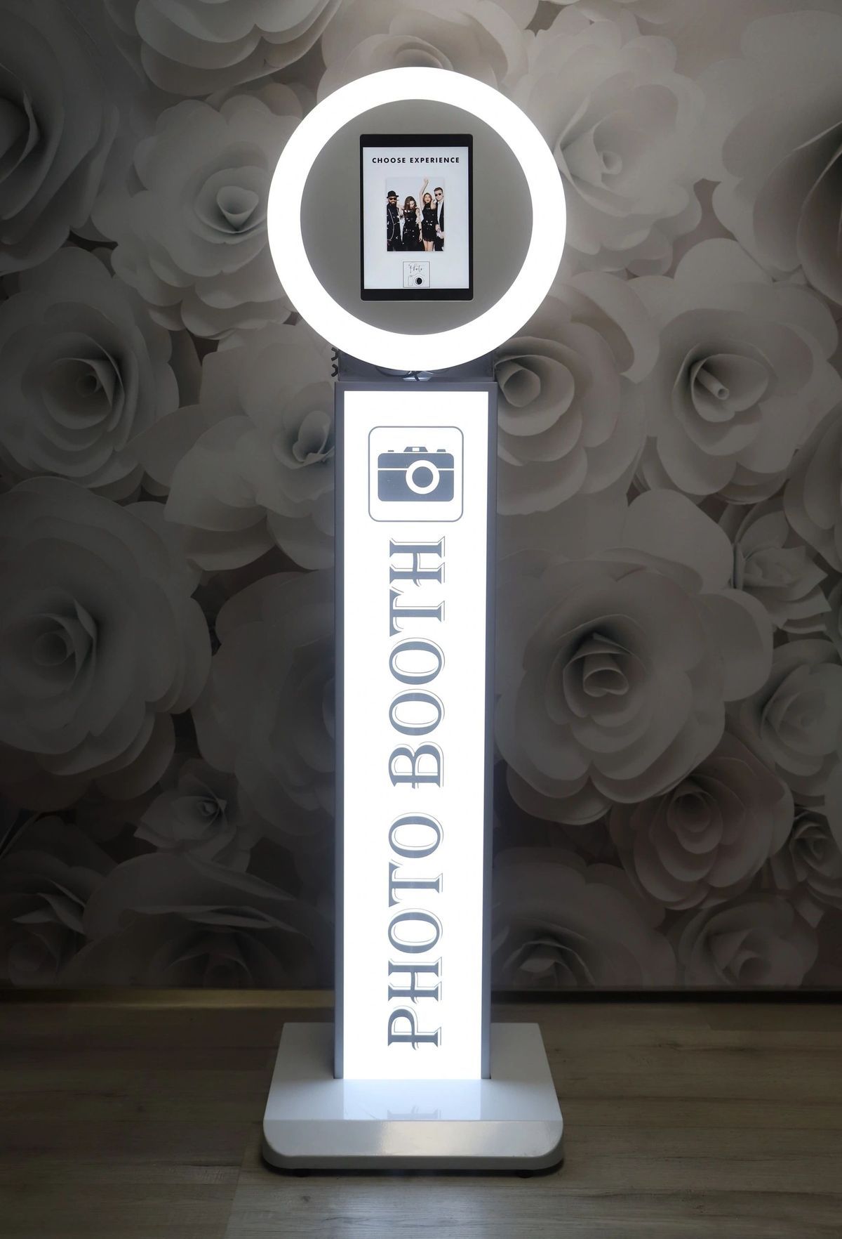 Three illuminated photo booth displays with white glowing signs stand against a floral wallpaper background.