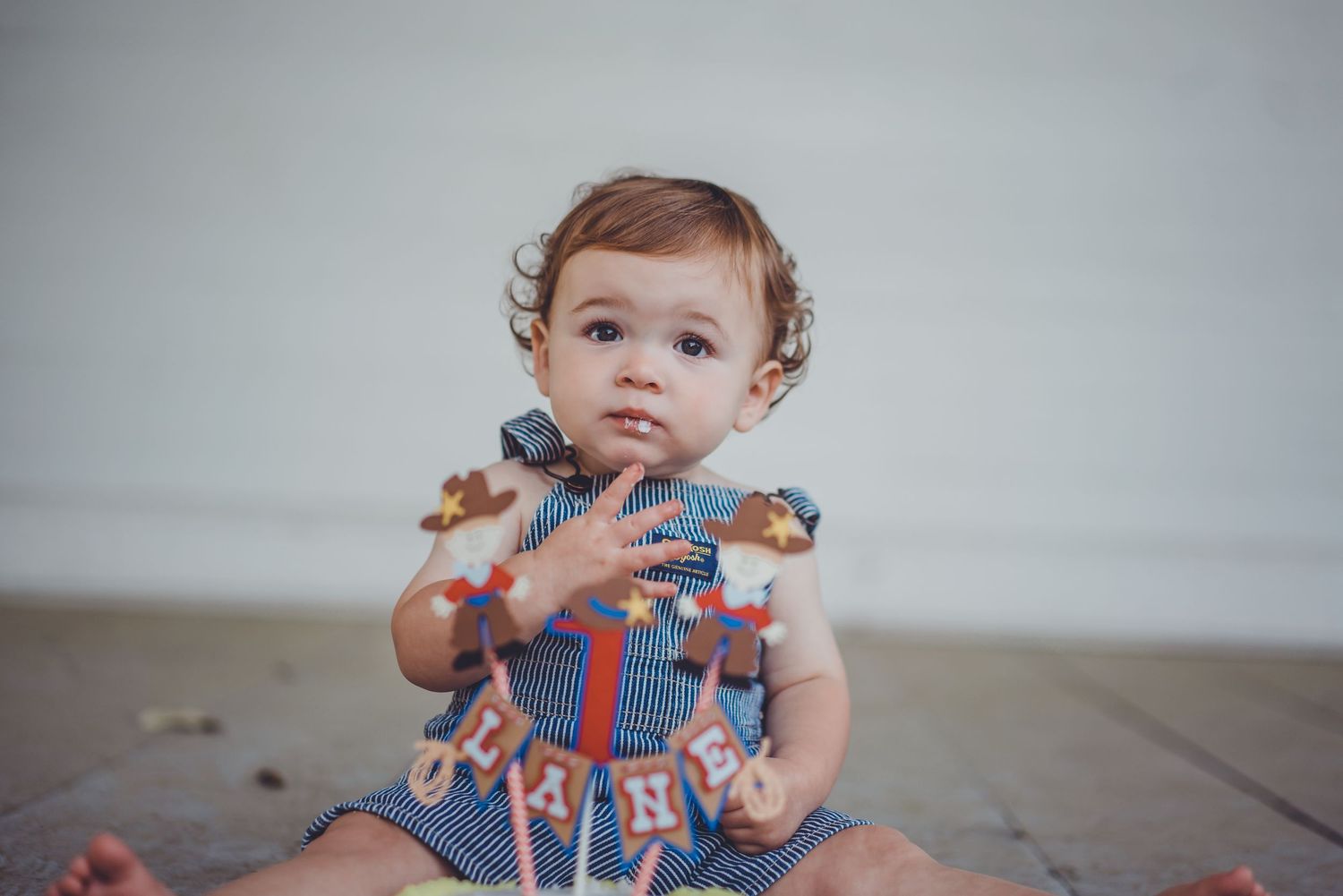 A baby in a patterned outfit sits on a wooden floor looking curious.