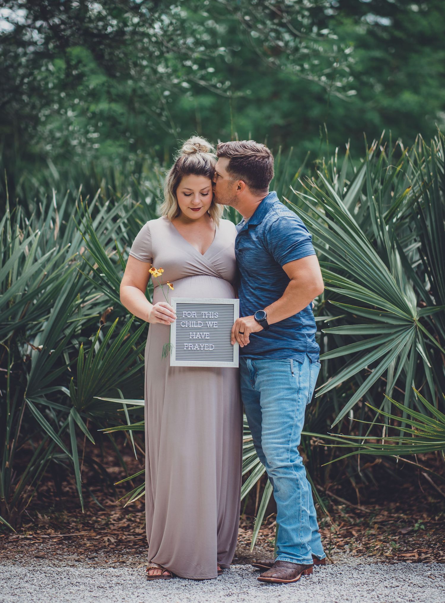 Couple poses with chalkboard sign surrounded by tropical plants and greenery.