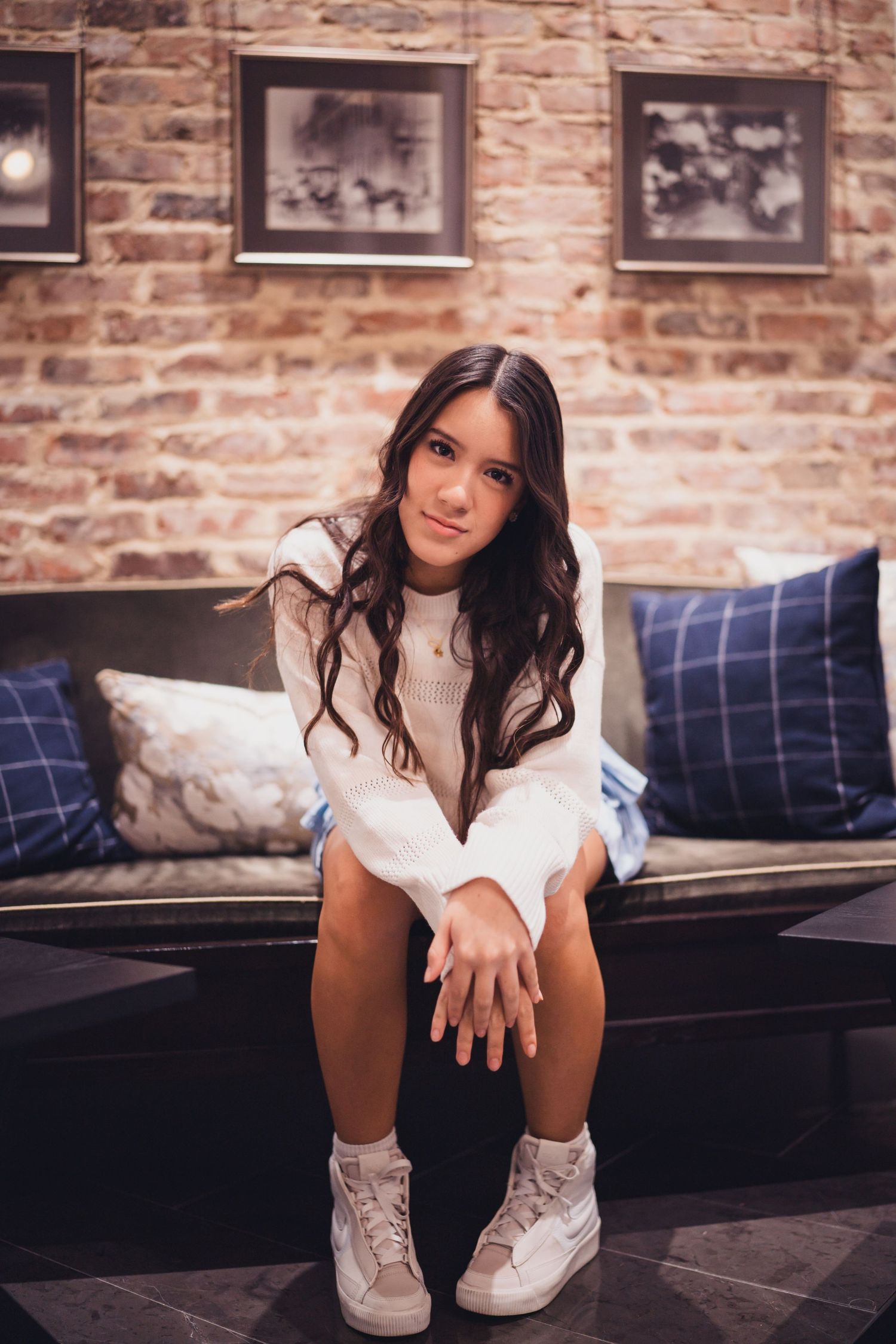 A person in a white sweater and sneakers sits casually on a couch against an exposed brick wall with framed photos.
