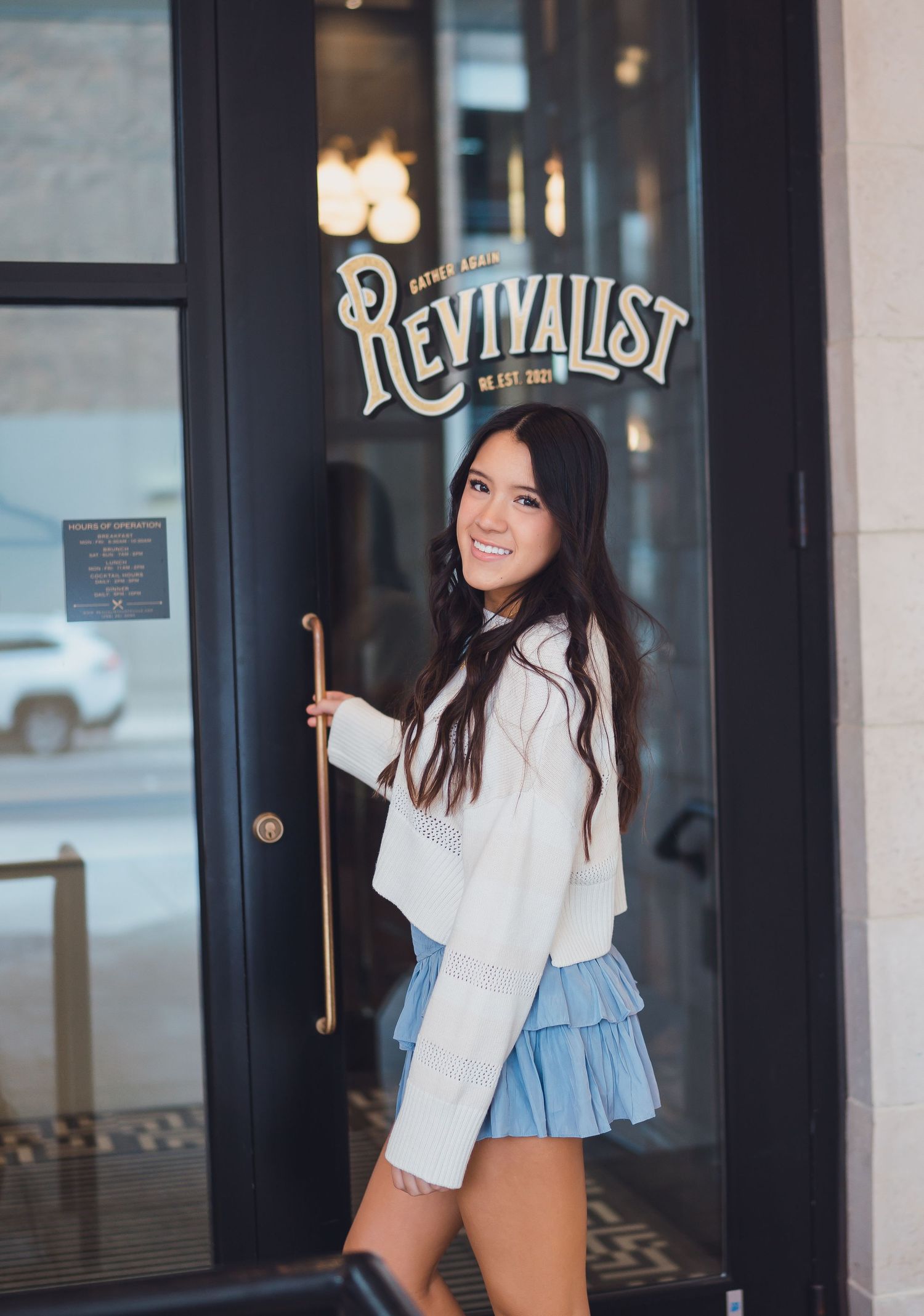 A person in a white blazer and denim shorts stands outside a storefront with black doors and illuminated signage.