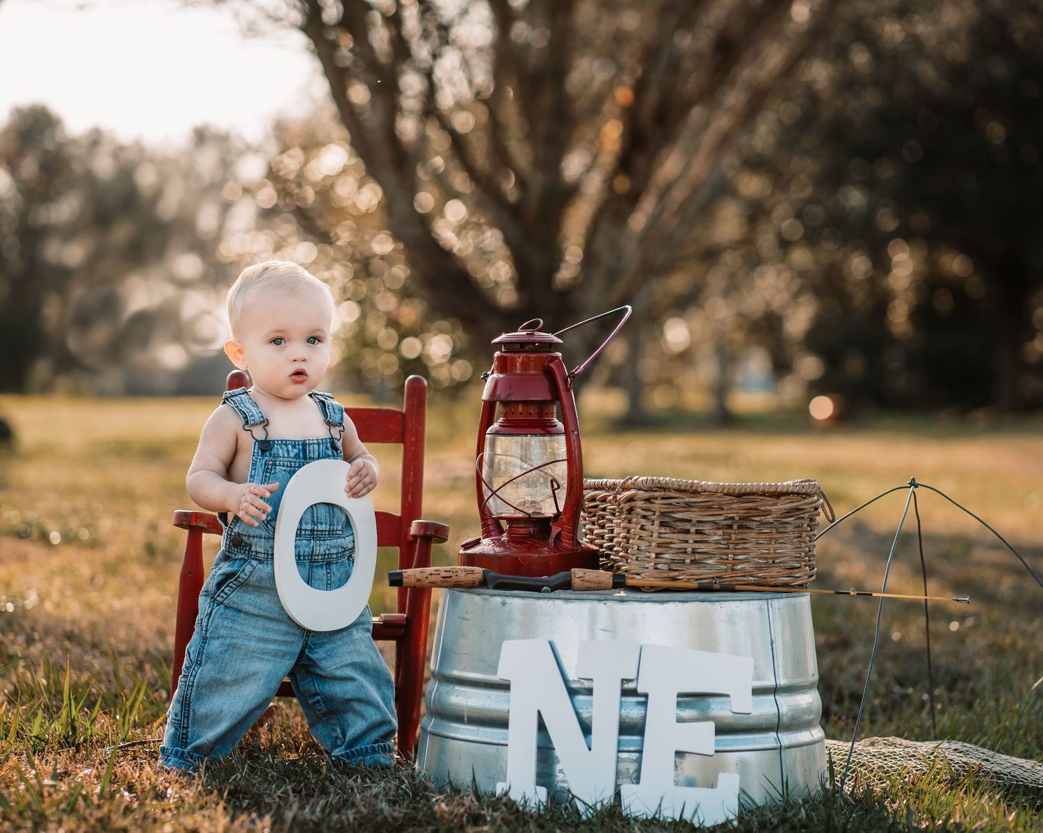 Toddler in overalls sits in red chair next to vintage lantern and ONE sign during sunset photoshoot.