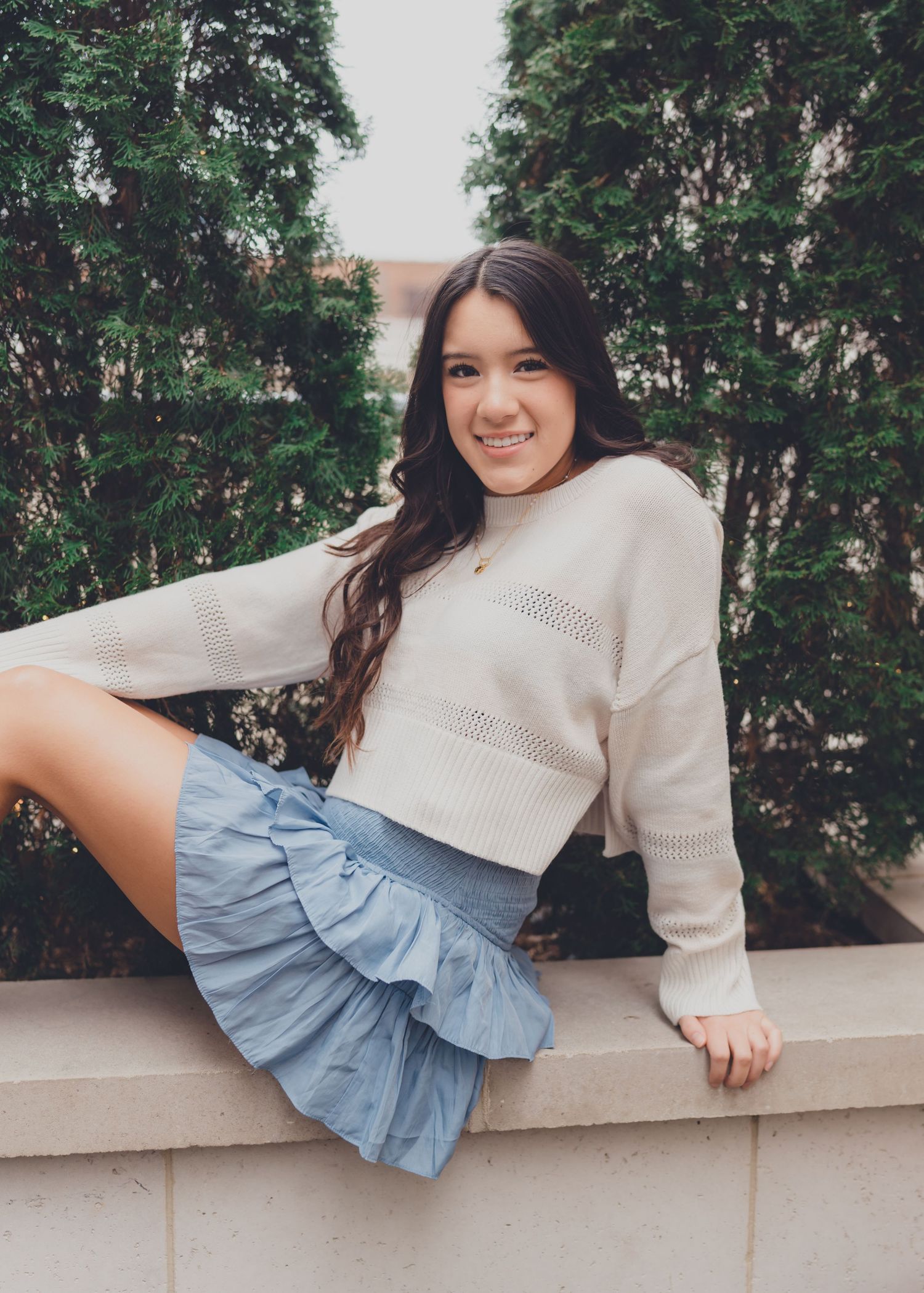 A person in a cream sweater and denim skirt poses playfully on a concrete ledge against green foliage.