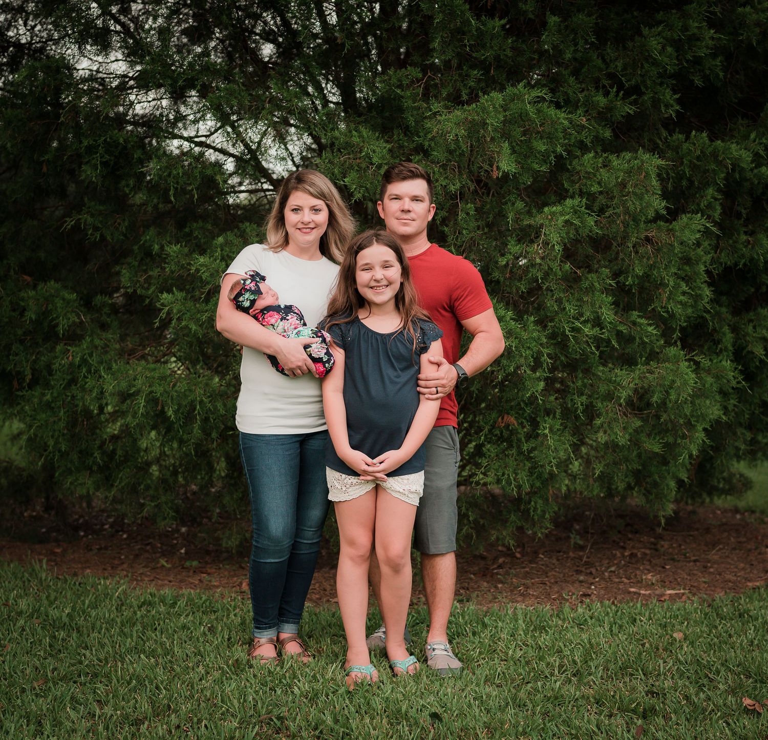 Family of three standing together outdoors against green evergreen background.