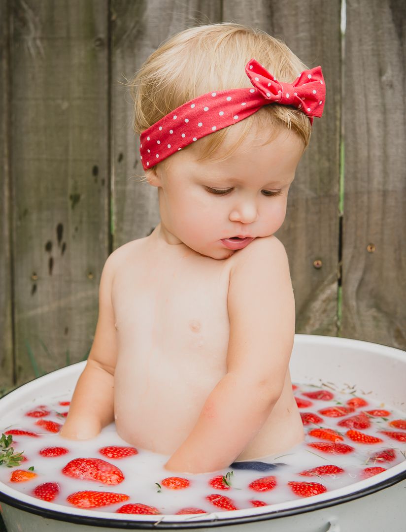 Baby in red polka dot headband sitting in milk bath with floating strawberries.