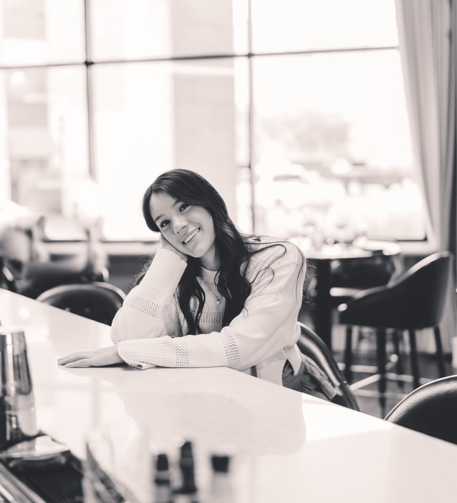 Black and white photo of someone sitting at a table by a window in a restaurant, resting their head on their hand.