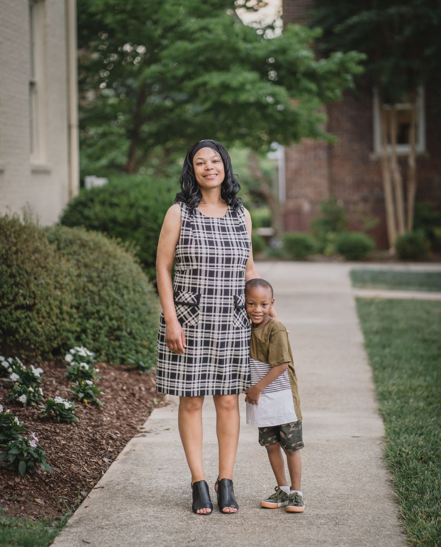 A woman in a plaid dress and child in brown clothing stand together on a walkway surrounded by landscaping.