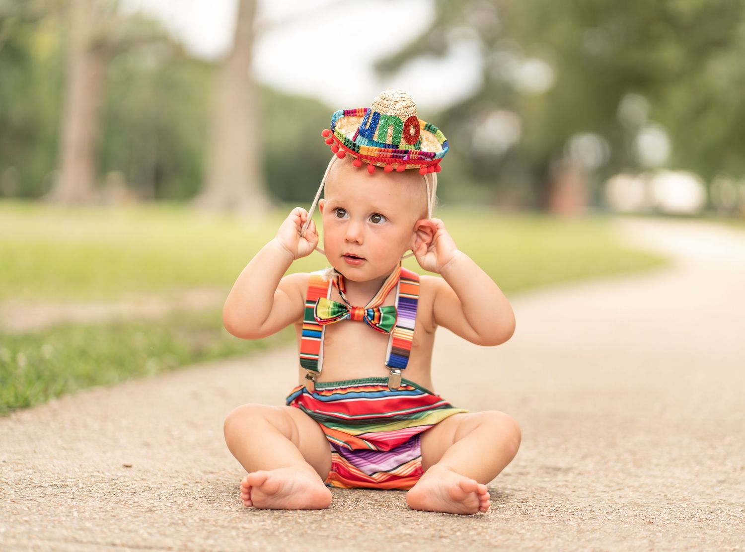 Baby wearing colorful striped outfit sits on path outdoors with festive hat and suspenders.