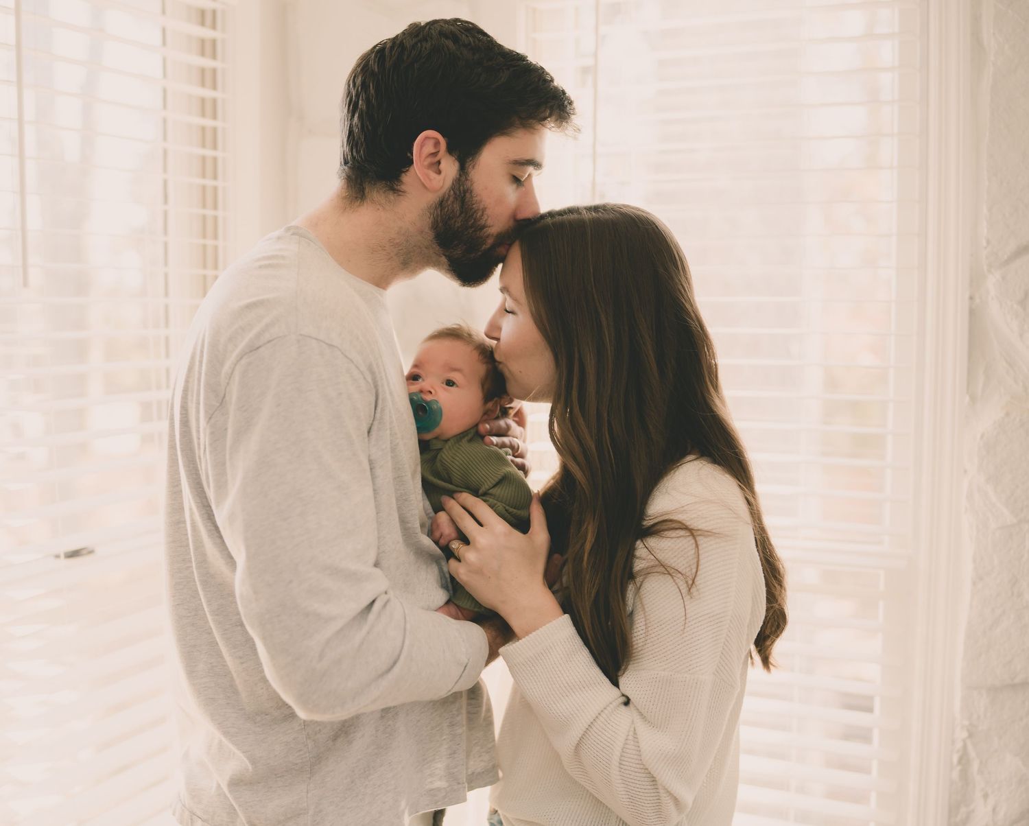 Parents share an intimate moment with their newborn baby by a sunlit window.