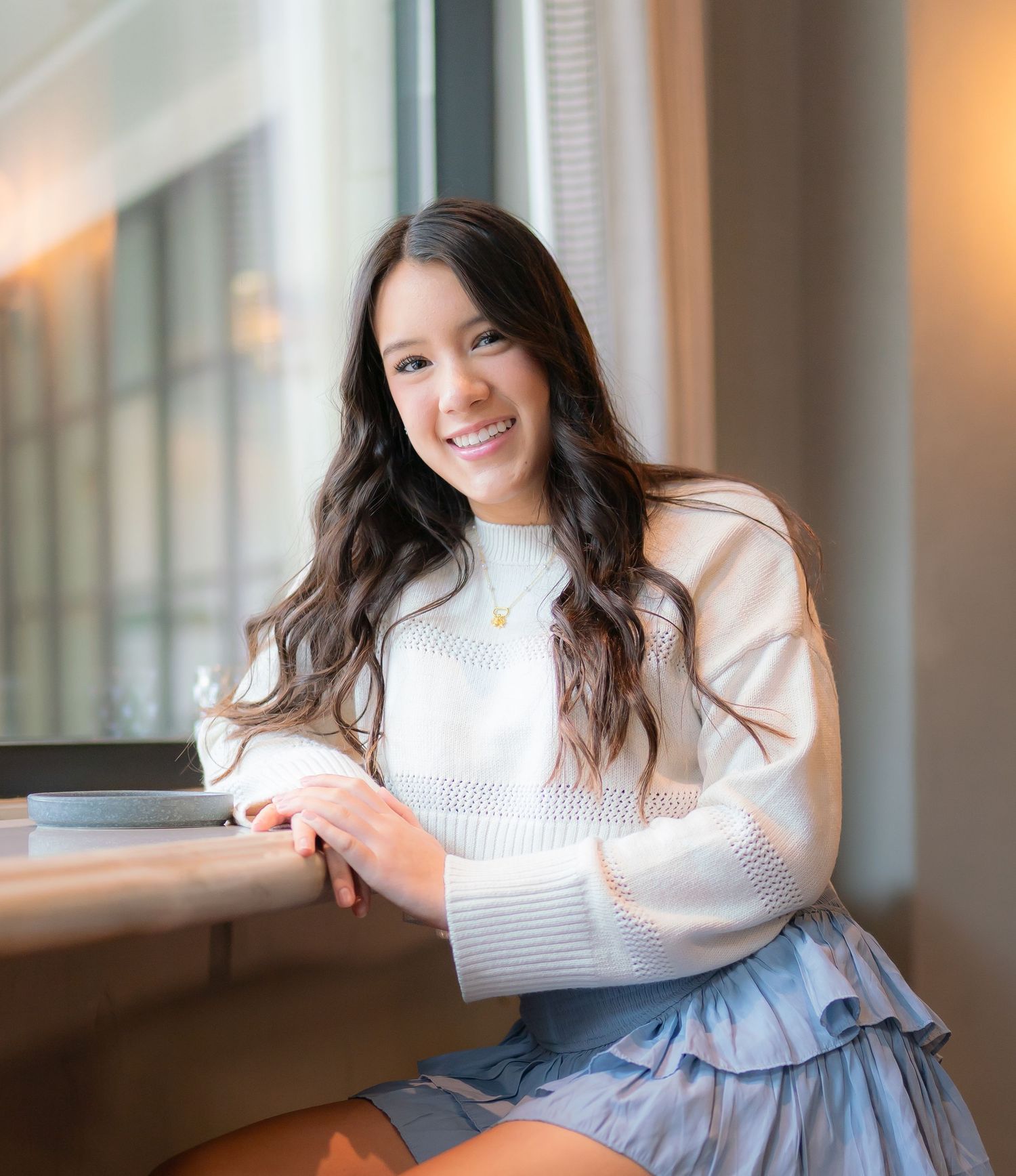 Person in white sweater and light blue skirt sits by window in warm interior lighting.