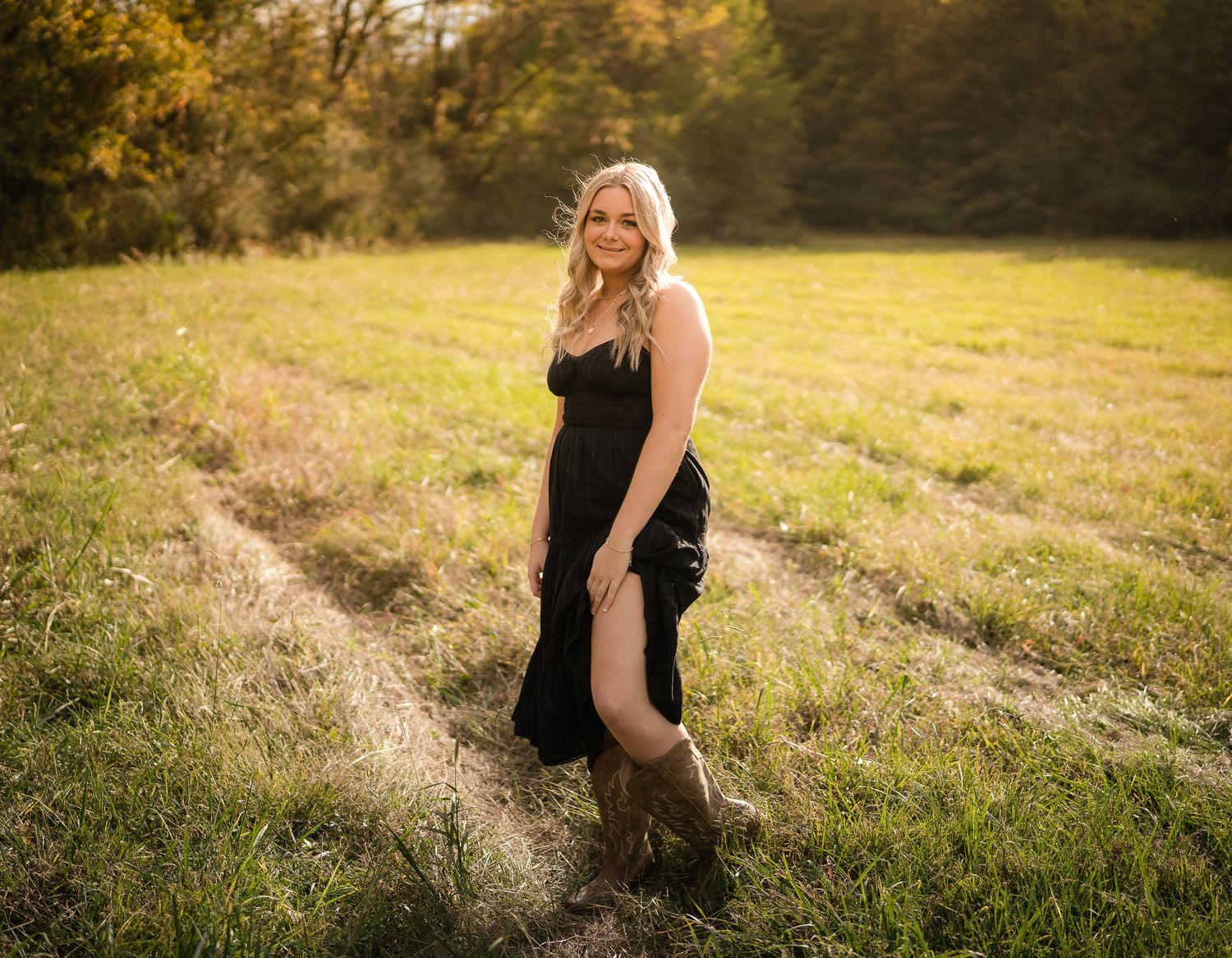 A figure in a black dress poses gracefully in a golden sunlit meadow during a sunset photoshoot session.