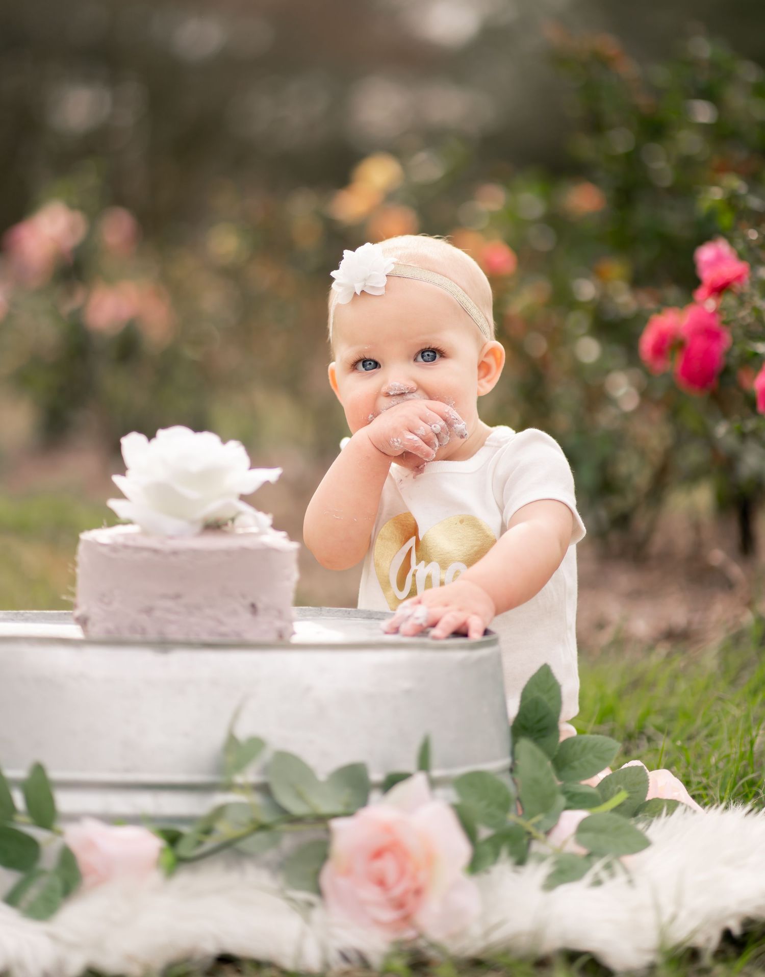 Cake smash photo session in a garden with roses and a white frosted cake on display.
