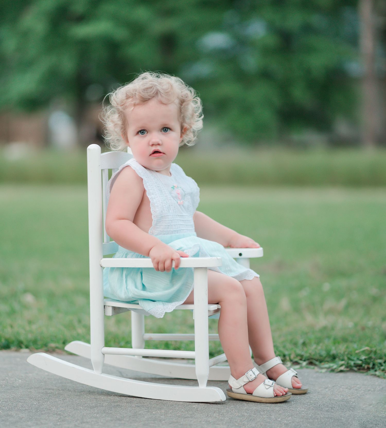 Small child in mint dress sits in white rocking chair outdoors.