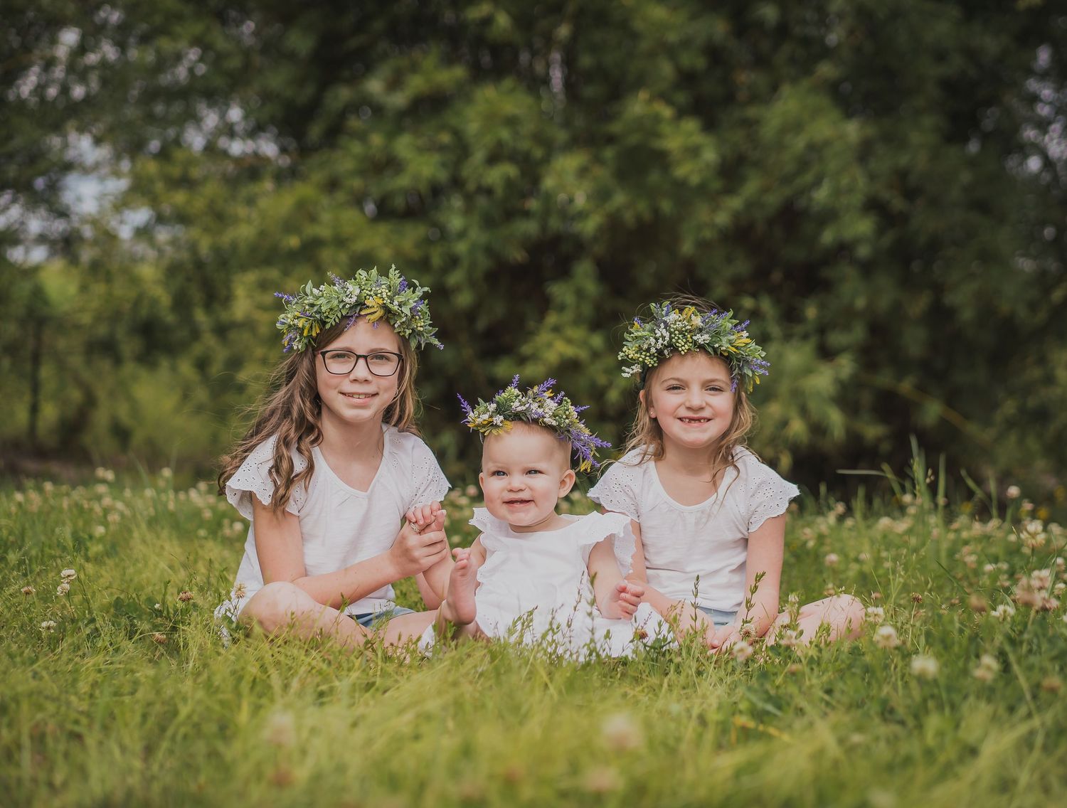 Three figures in white dresses adorned with floral crowns pose together in a wildflower field.