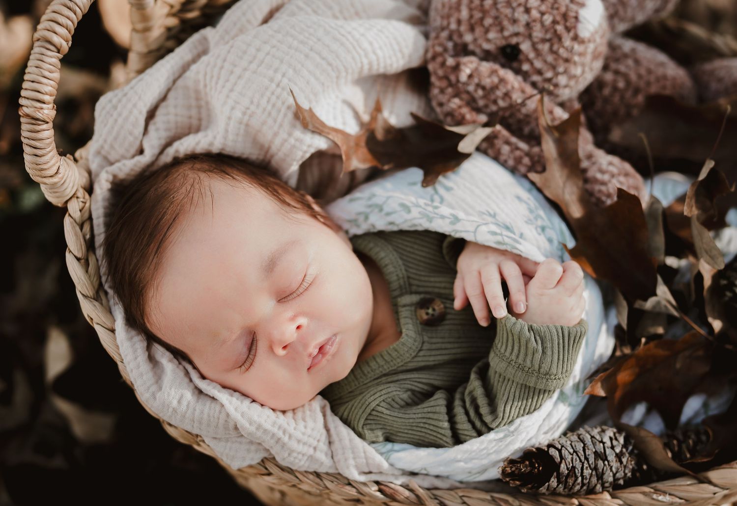 A sleeping baby in a white knit hat lies in a woven basket surrounded by autumn leaves and natural textures.