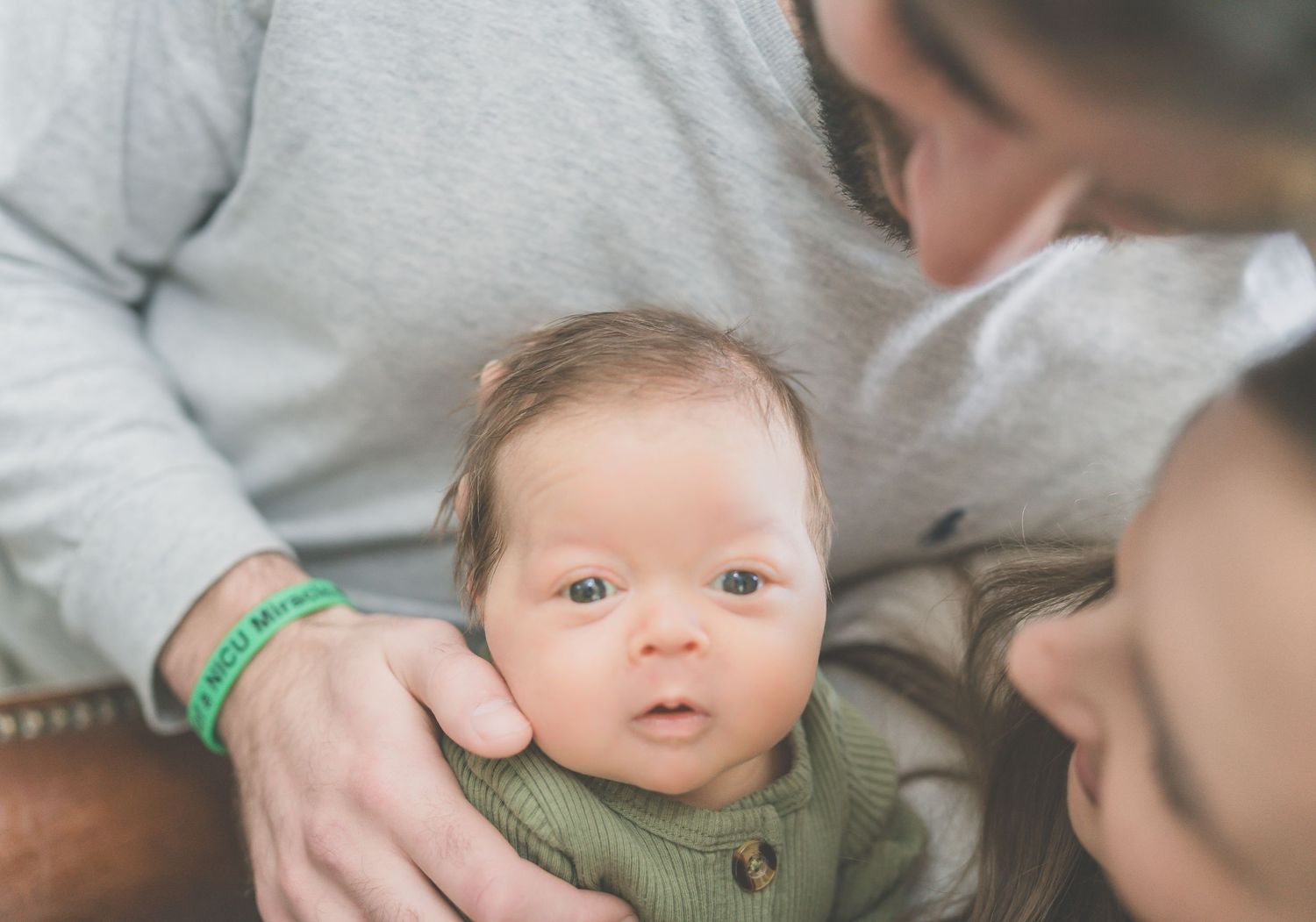 A baby in a green knit sweater smiles while being held by someone in a light-colored shirt.