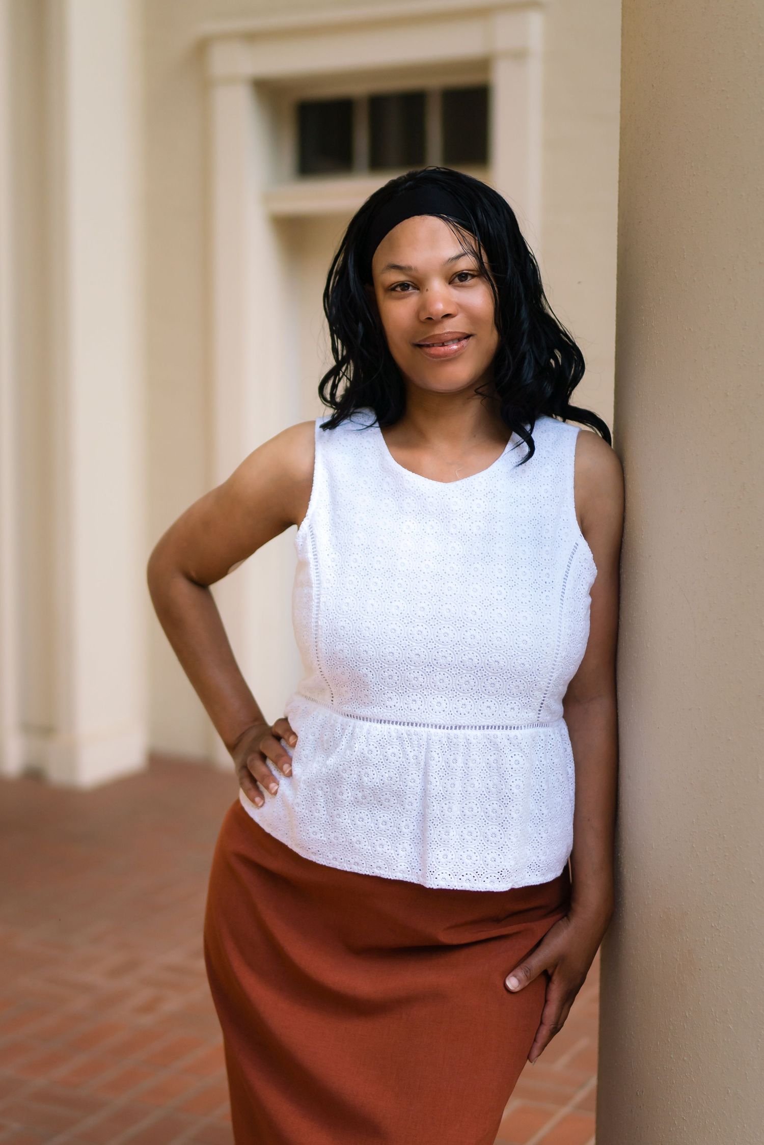 A person wearing a white sleeveless peplum top and brown pencil skirt poses confidently against a light colored wall.