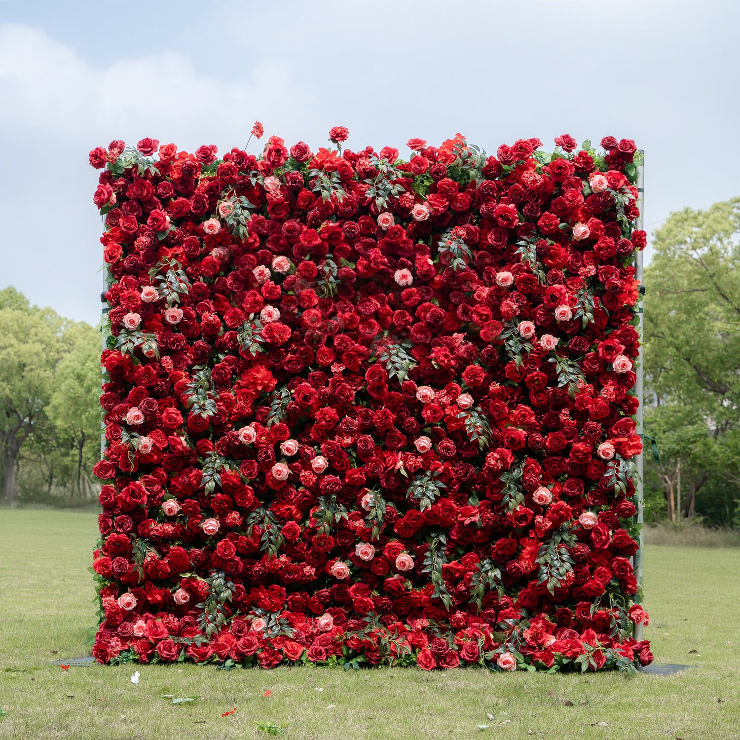 A dramatic wall of deep red roses and floral arrangements displayed outdoors on a lawn.