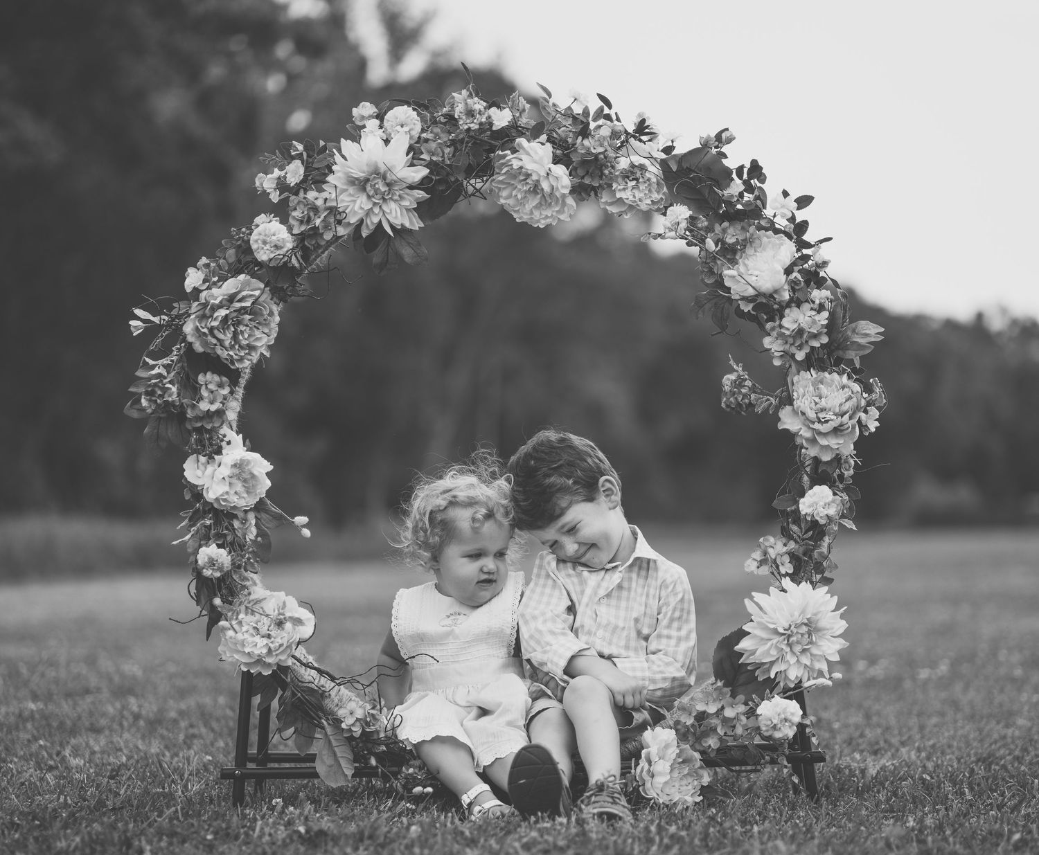 Black and white portrait of children sitting under floral arch outdoors.