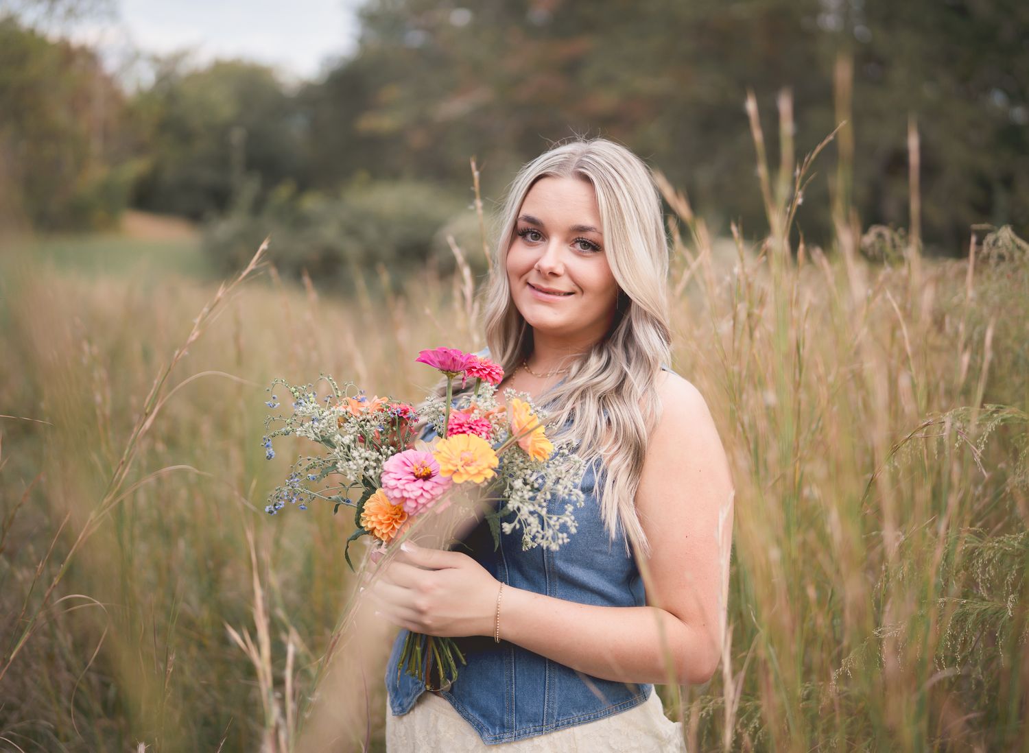 A woman in a denim dress holds a vibrant bouquet of wildflowers while standing in a golden field at sunset.