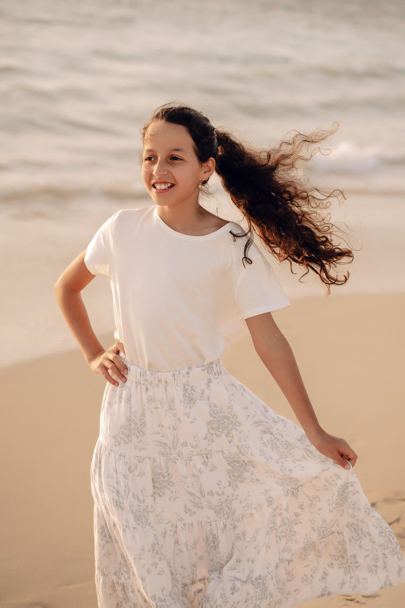 A girl in a white floral dress spins and dances on the beach in golden sunlight.