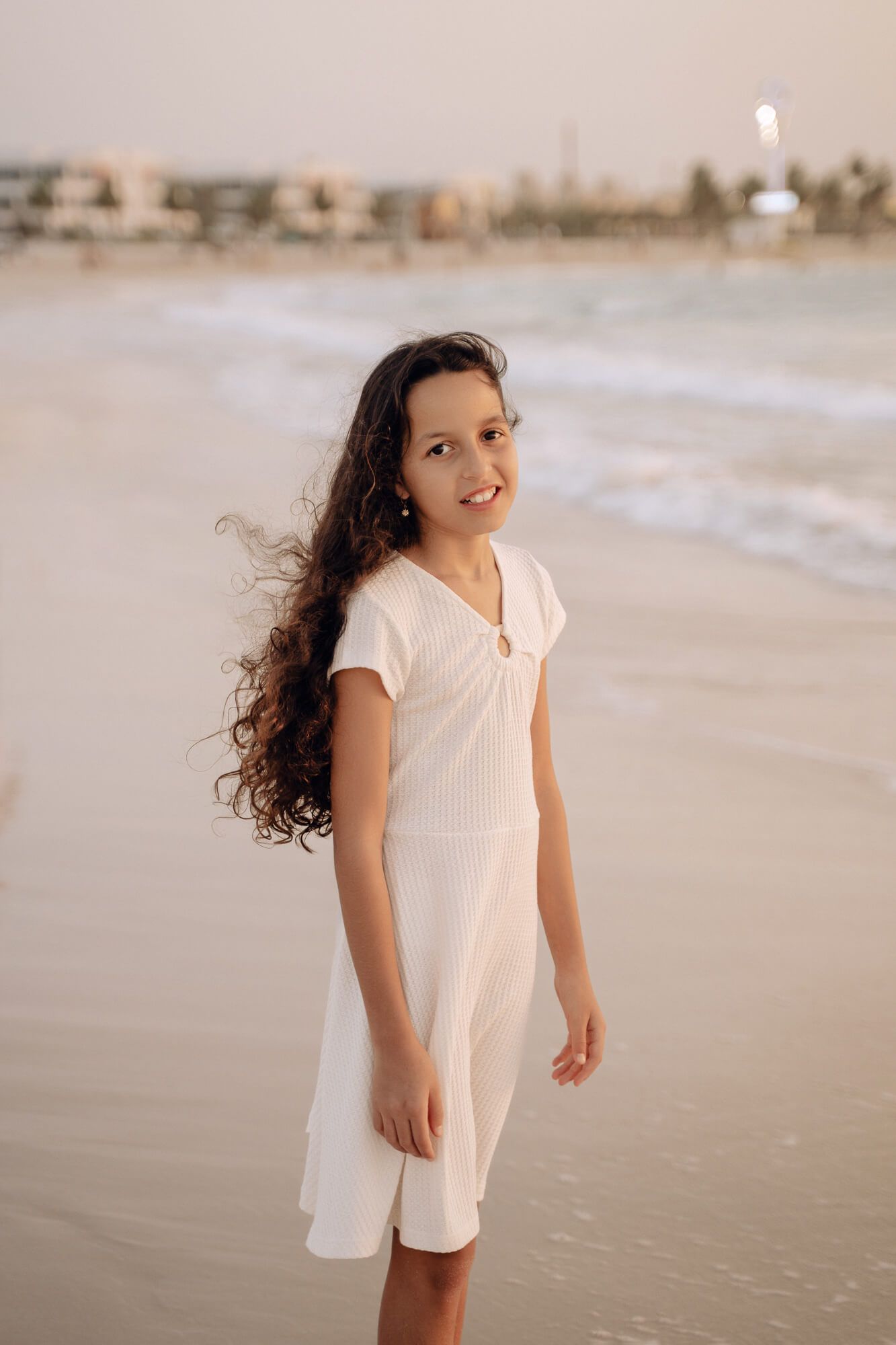 A person in a white dress poses on the beach during golden hour with cityscape in background.