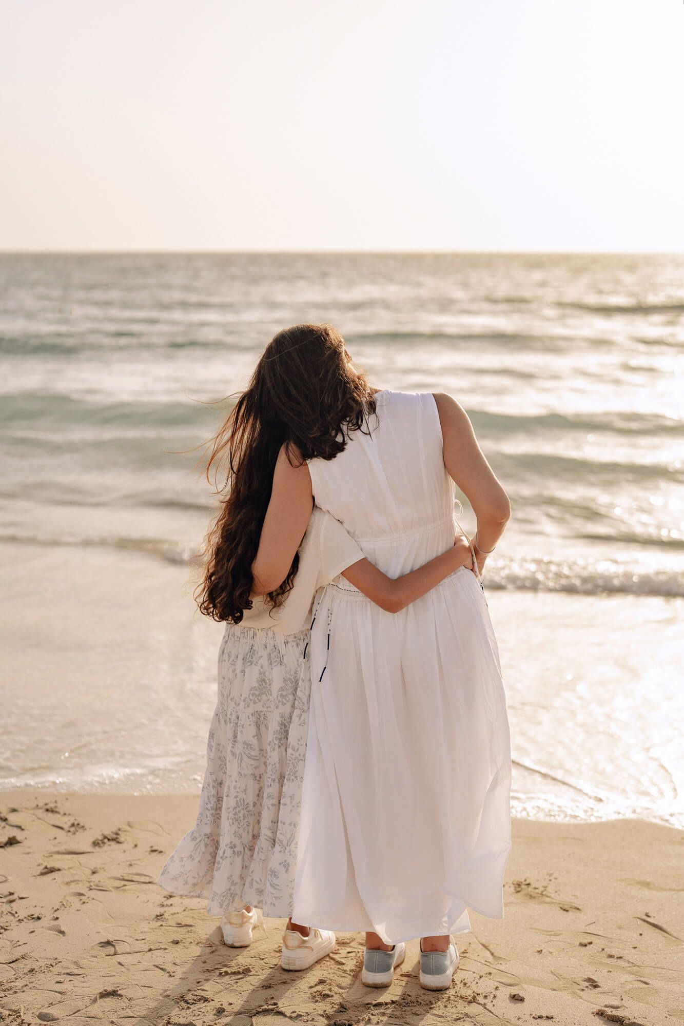 Two people in white dresses embrace while looking at the ocean waves during sunset.