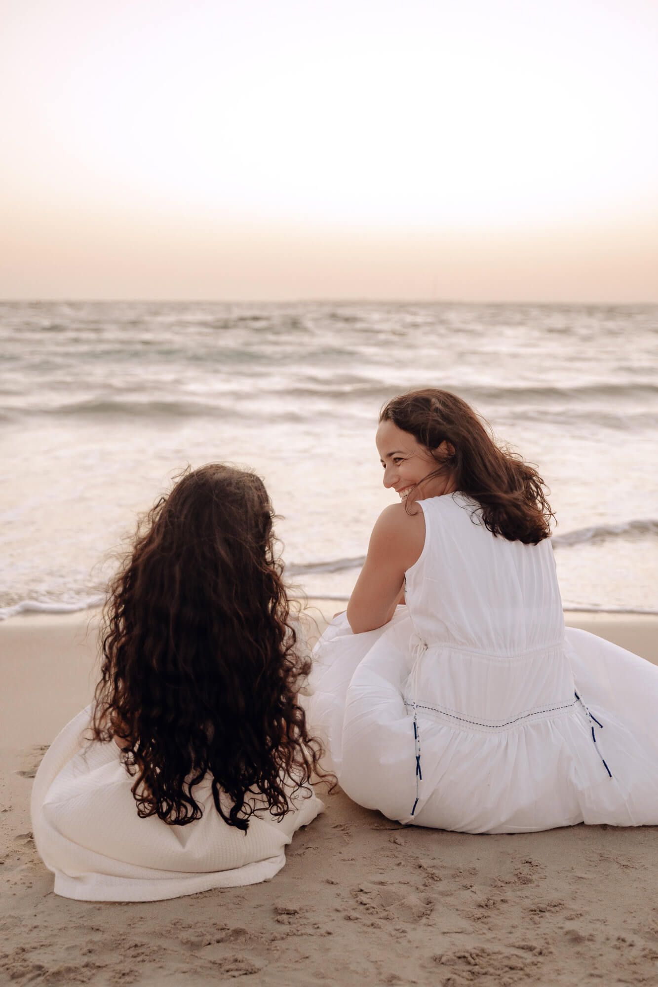 Two people in white dresses sitting together on beach watching ocean waves at sunset.