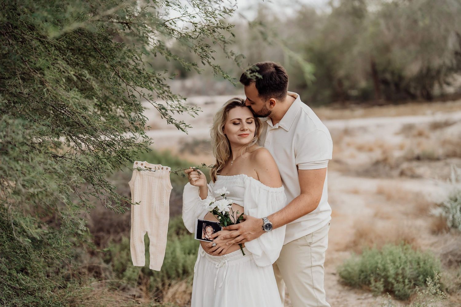 Intimate maternity photo of couple in white holding baby clothes and flowers.