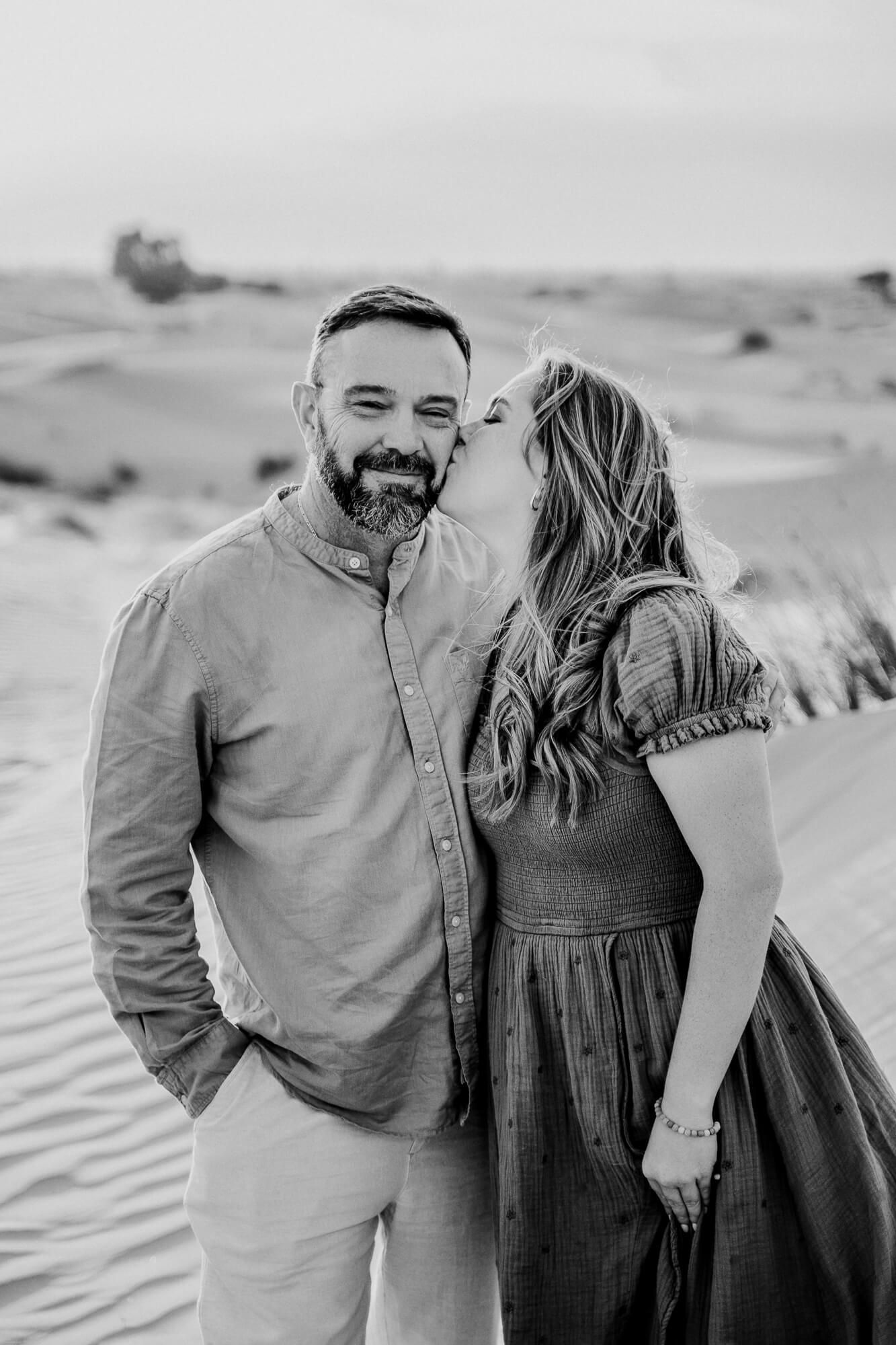 Black and white portrait of a couple sharing an affectionate moment in a desert landscape.