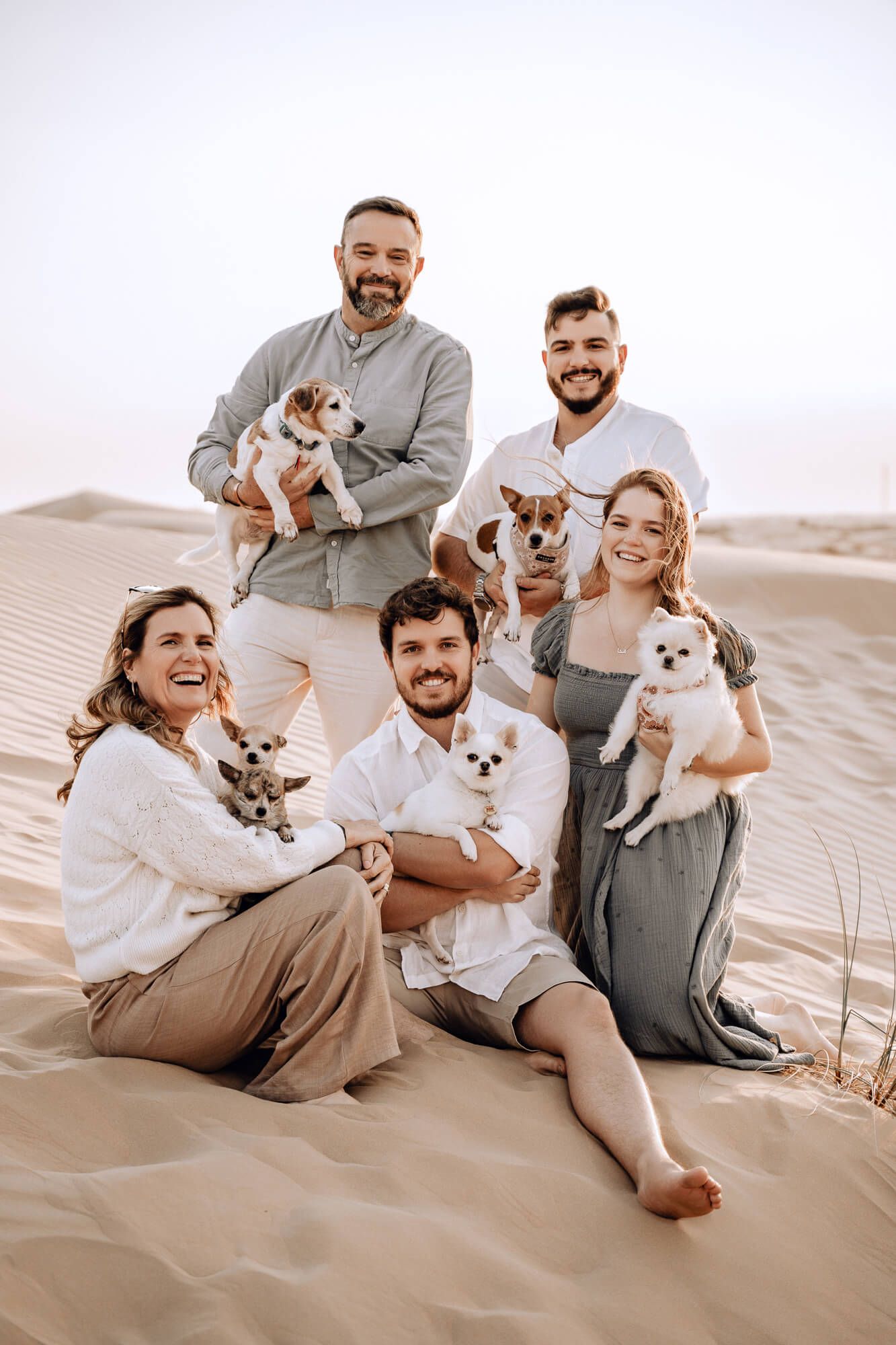 Group portrait of five people with their small dogs sitting together on desert sand dunes.