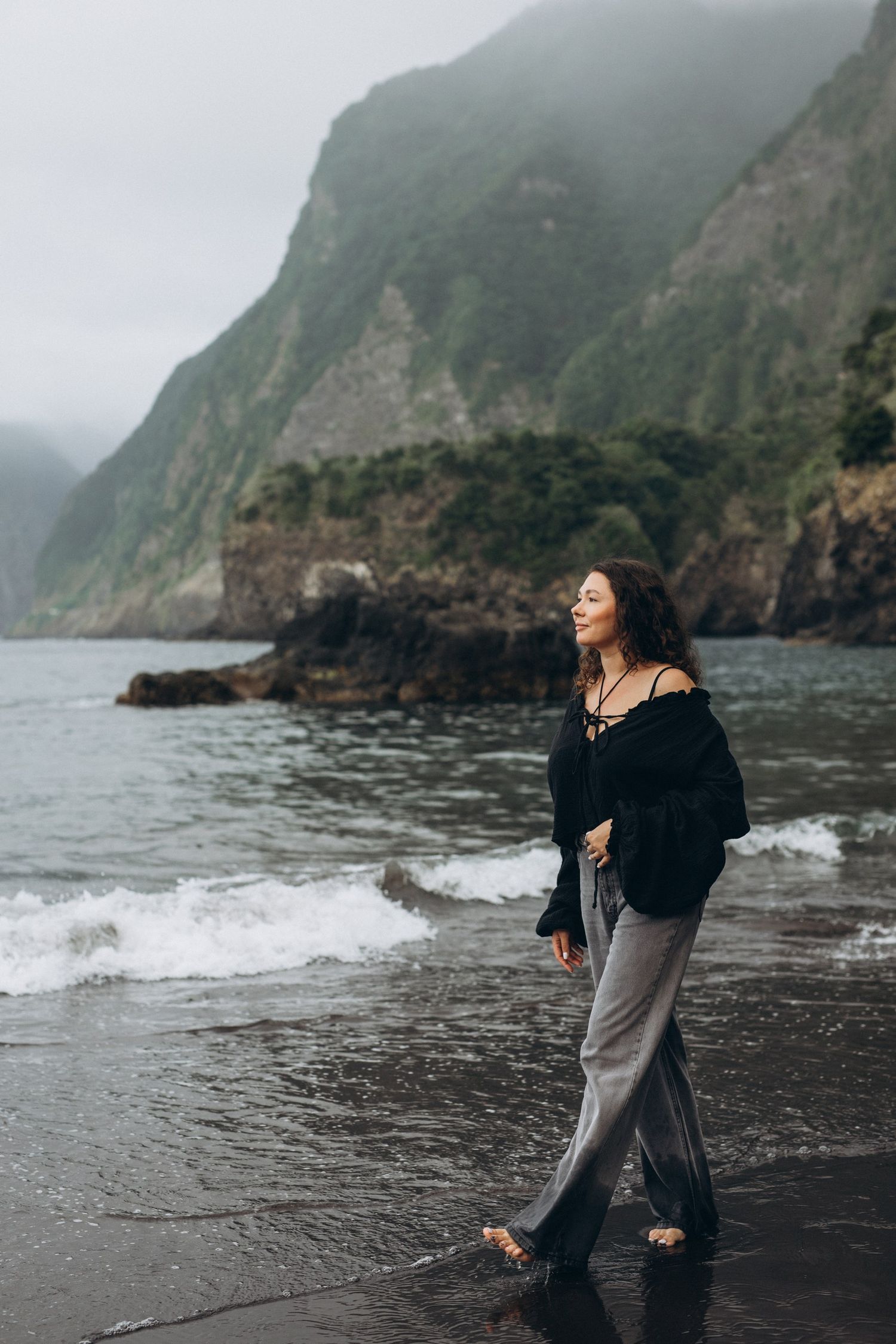 A misty seascape shows rocky cliffs descending into a dark beach where a person stands along the shoreline.