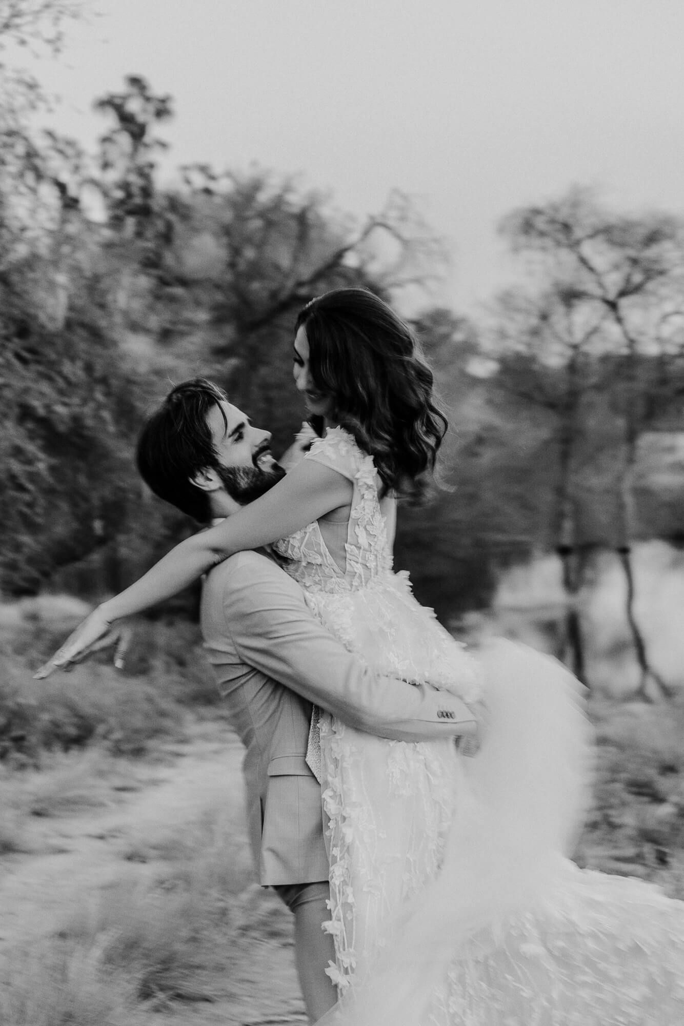 A couple in wedding attire hugs and spins joyfully in a scenic outdoor setting, captured in black and white photography.