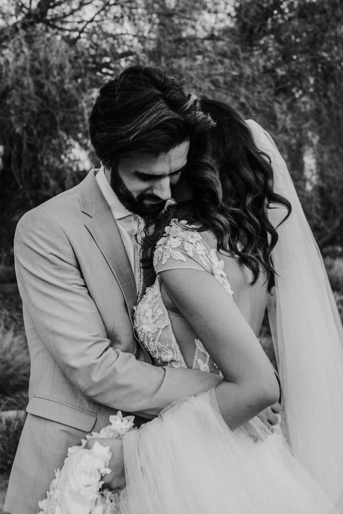 Black and white wedding photo shows a romantic embrace between newlyweds, with the bride in lace and groom in light suit.