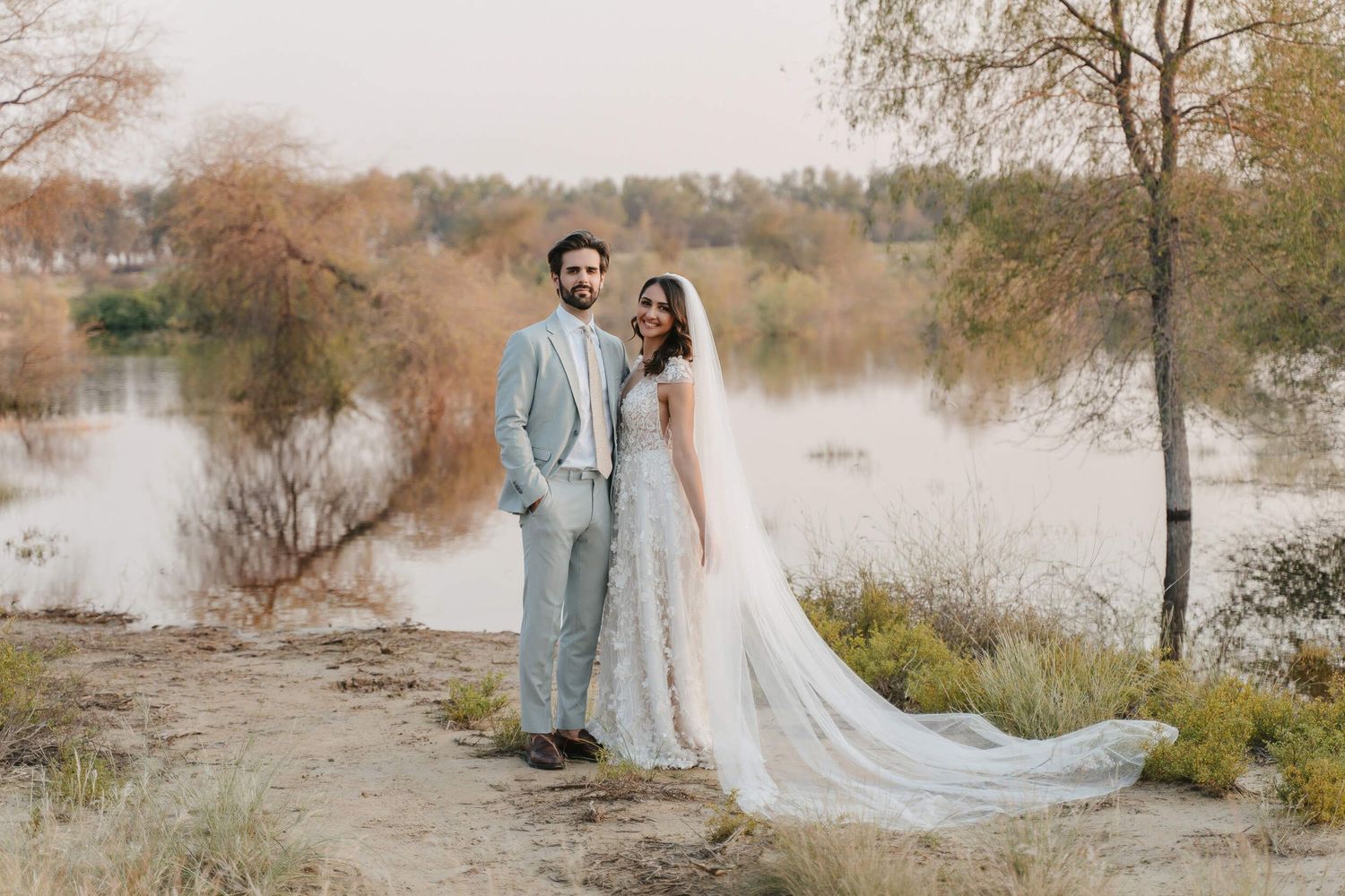 A couple in wedding attire poses together near a serene autumn lakeside with bare trees in the background.