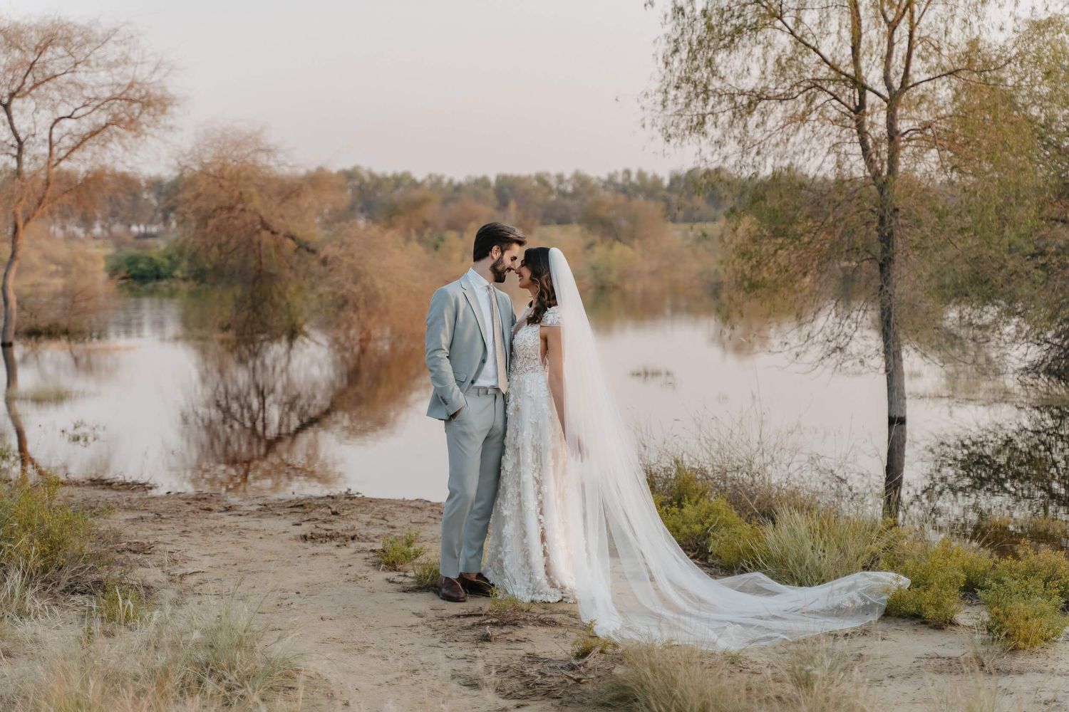 A romantic wedding photo by a lake with fall foliage, featuring a couple in gray suit and flowing white gown.