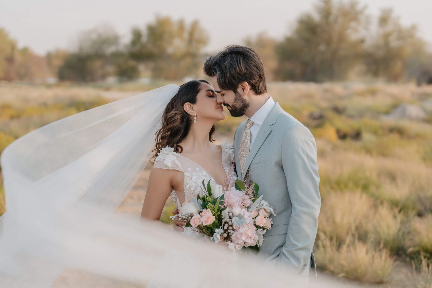 A romantic desert wedding shoot with a bride and groom kissing in golden sunlit fields during sunset.