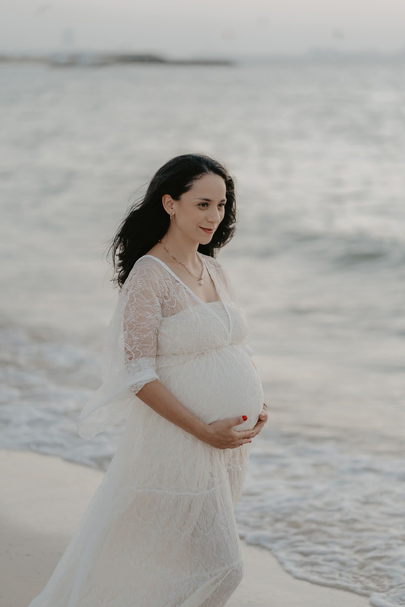 An expectant mother wearing a flowing white lace dress poses gracefully by the ocean shore during a maternity photoshoot.