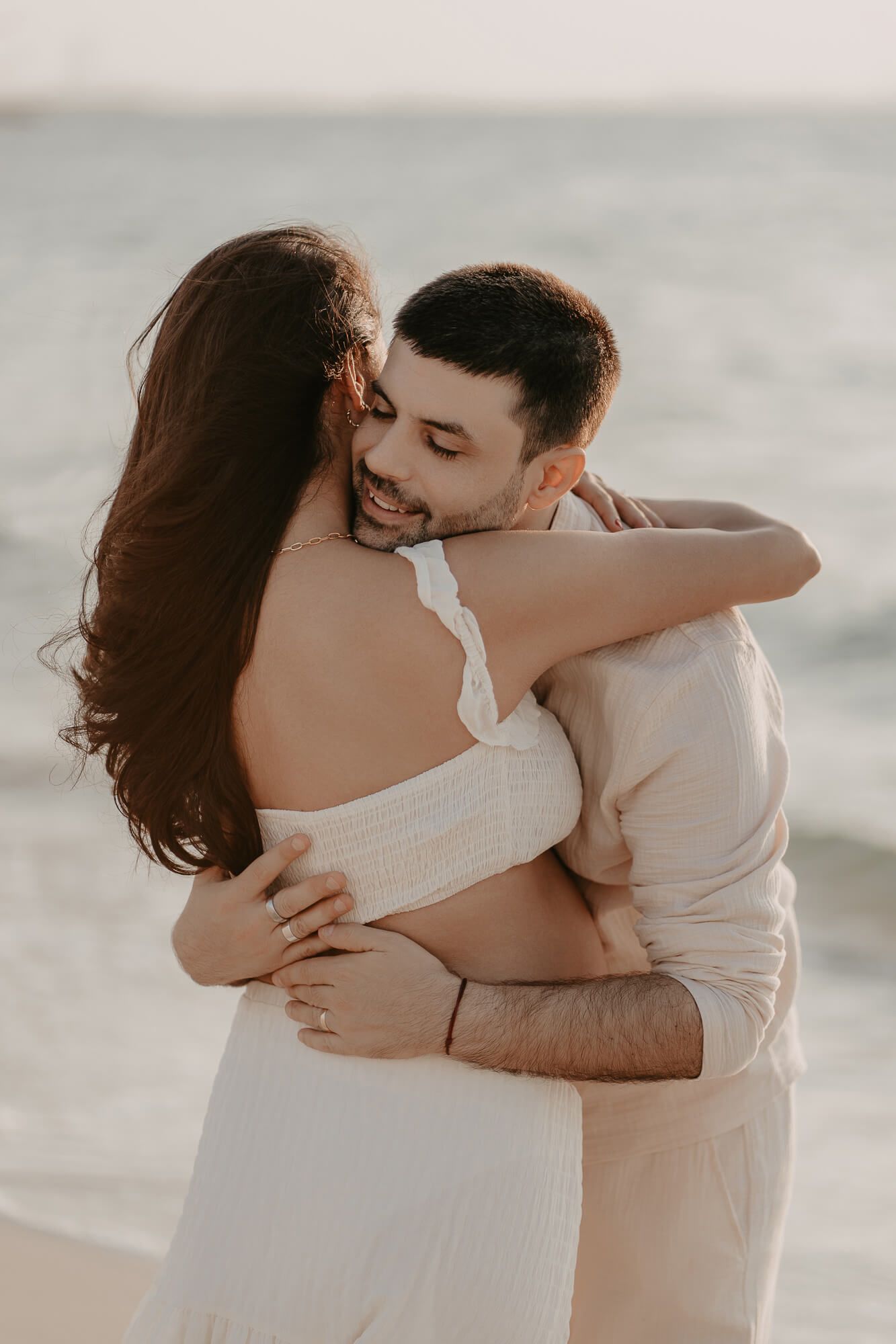 A romantic couple in white embraces each other tenderly by the water in a dreamy seaside photoshoot.