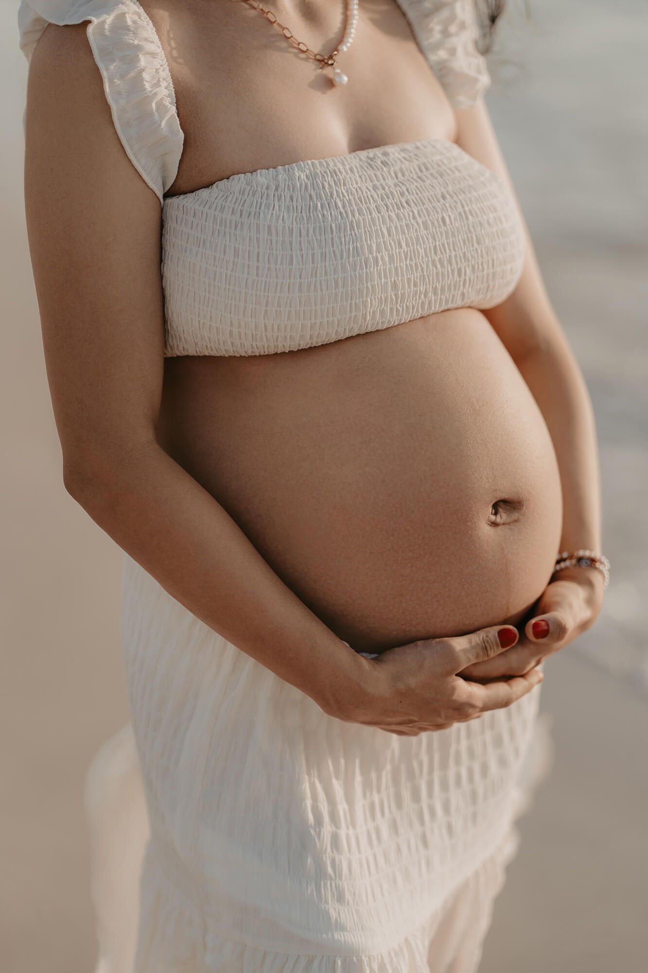 A pregnant woman in a white smocked top and skirt cradles her belly in a soft, ethereal maternity photo.