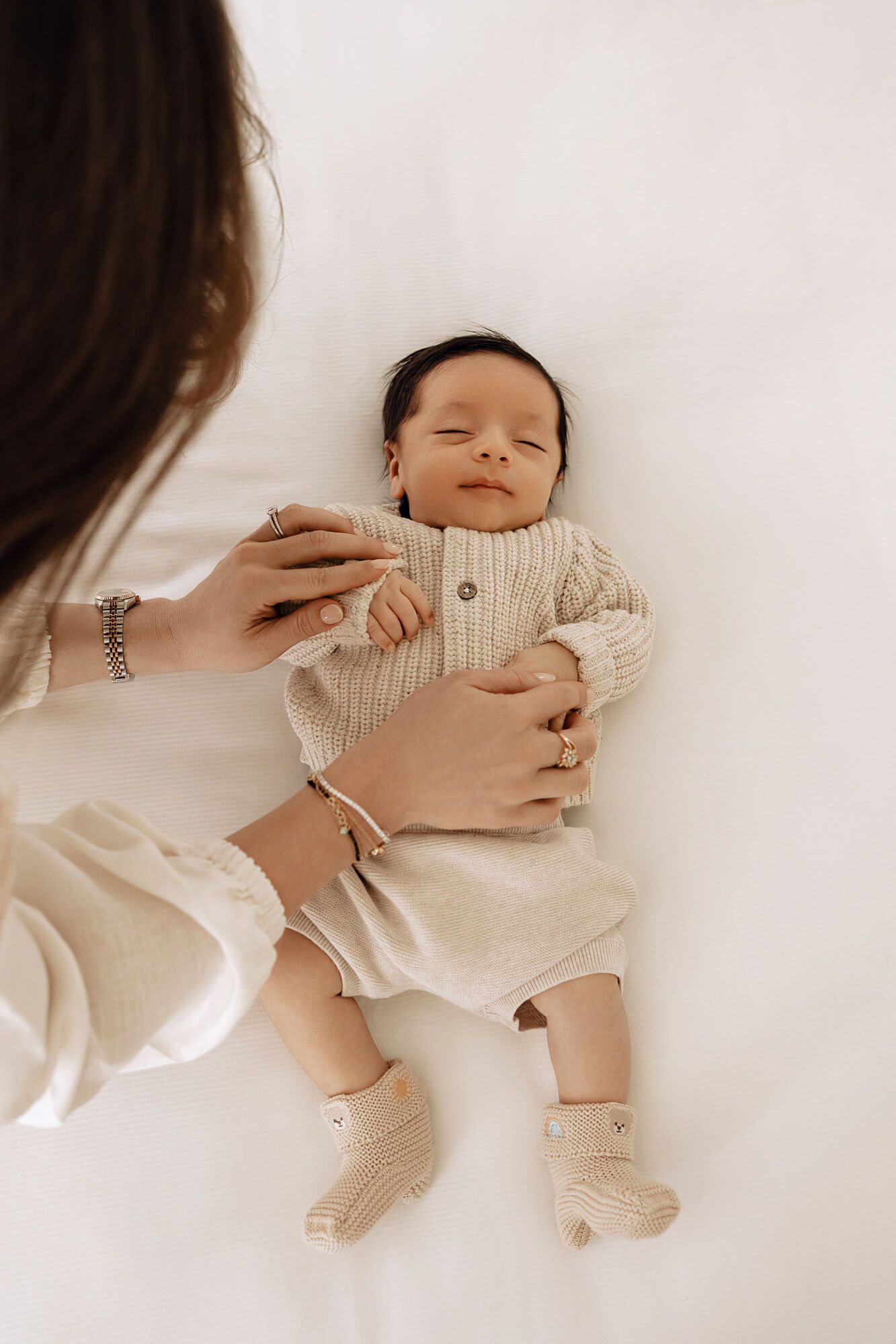 Baby in cream knit outfit lying on white bedding during lifestyle photoshoot.
