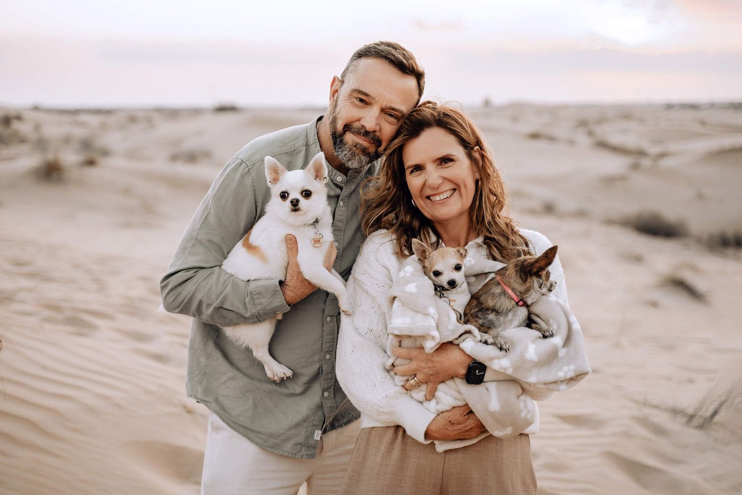 A couple poses with their small chihuahua dog in a desert landscape during sunset.