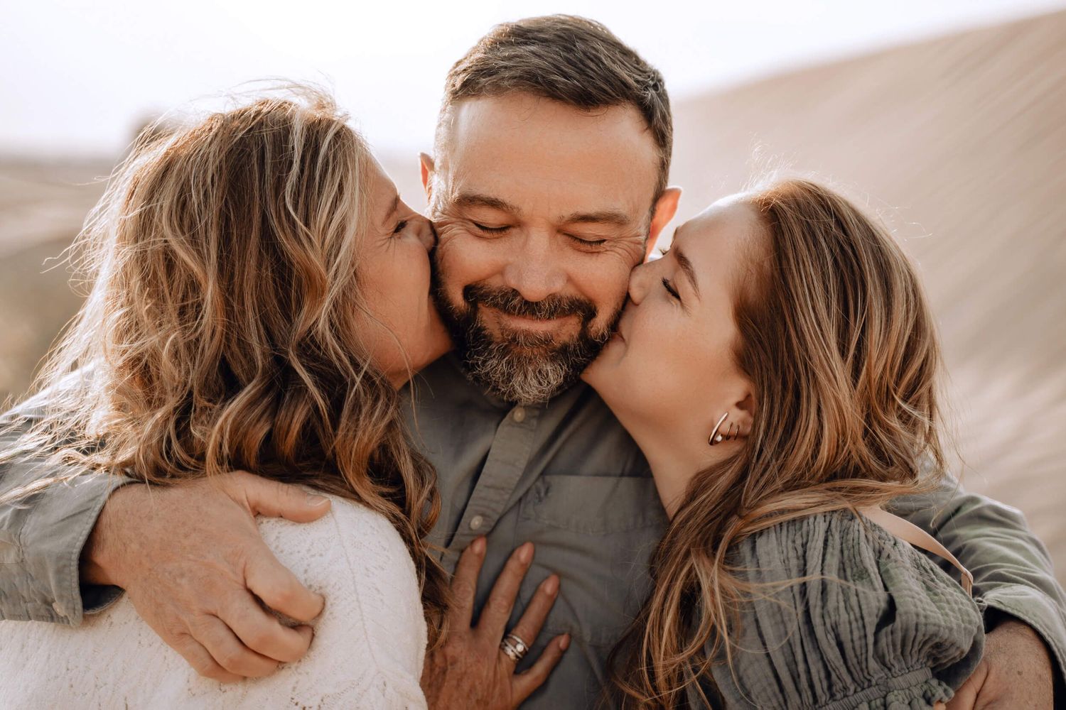 Candid moment of three people sharing affectionate kisses against sandy desert backdrop.