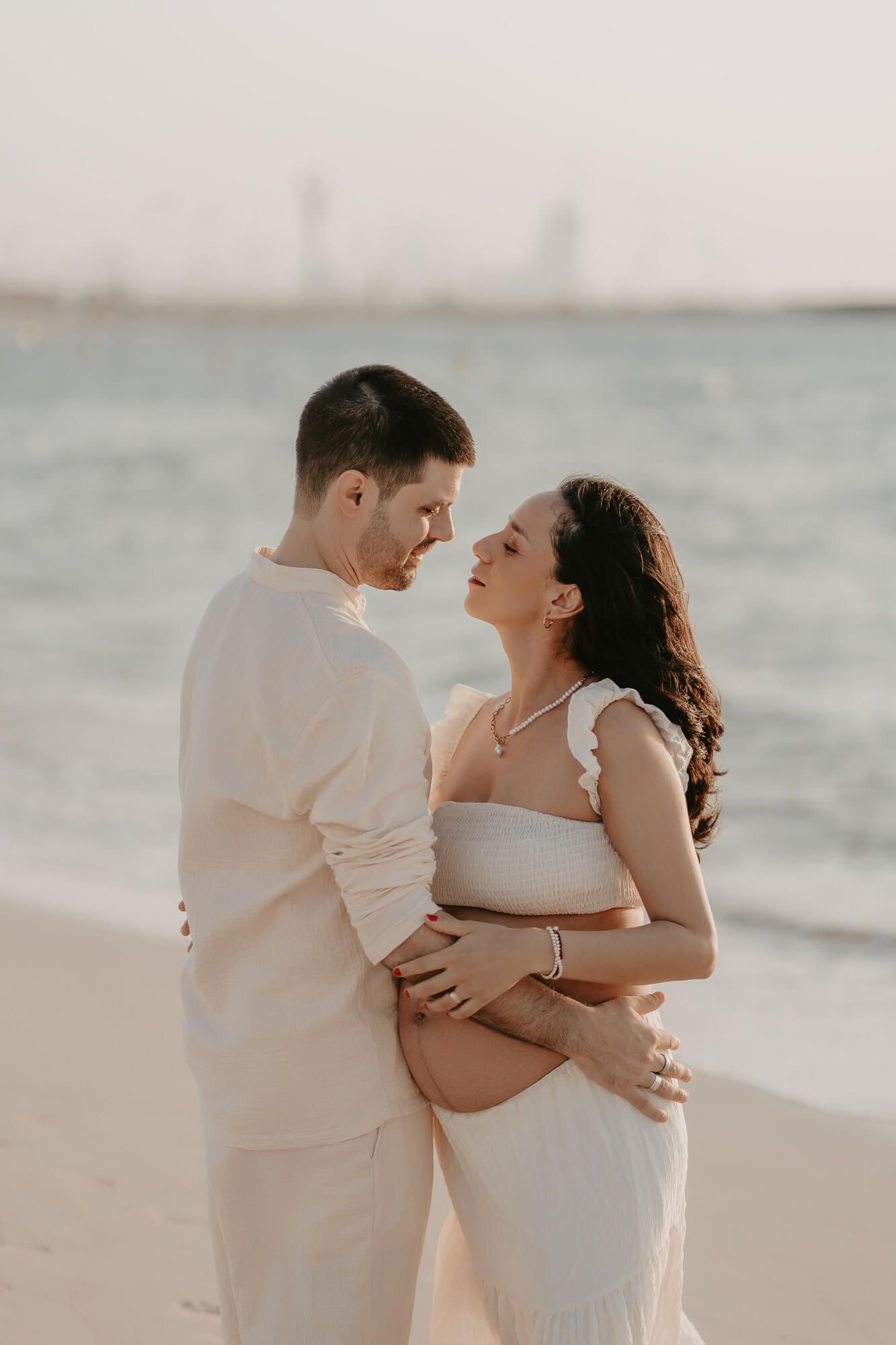 A romantic couple embraces on the beach at sunset wearing light colored clothing against a misty ocean backdrop.