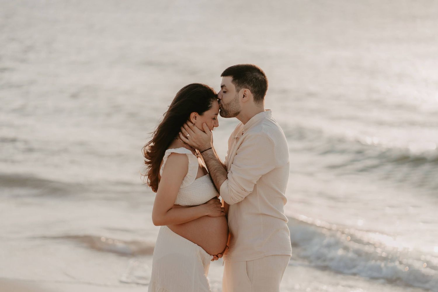 Expecting couple embraces on a serene beach during sunset maternity photoshoot.